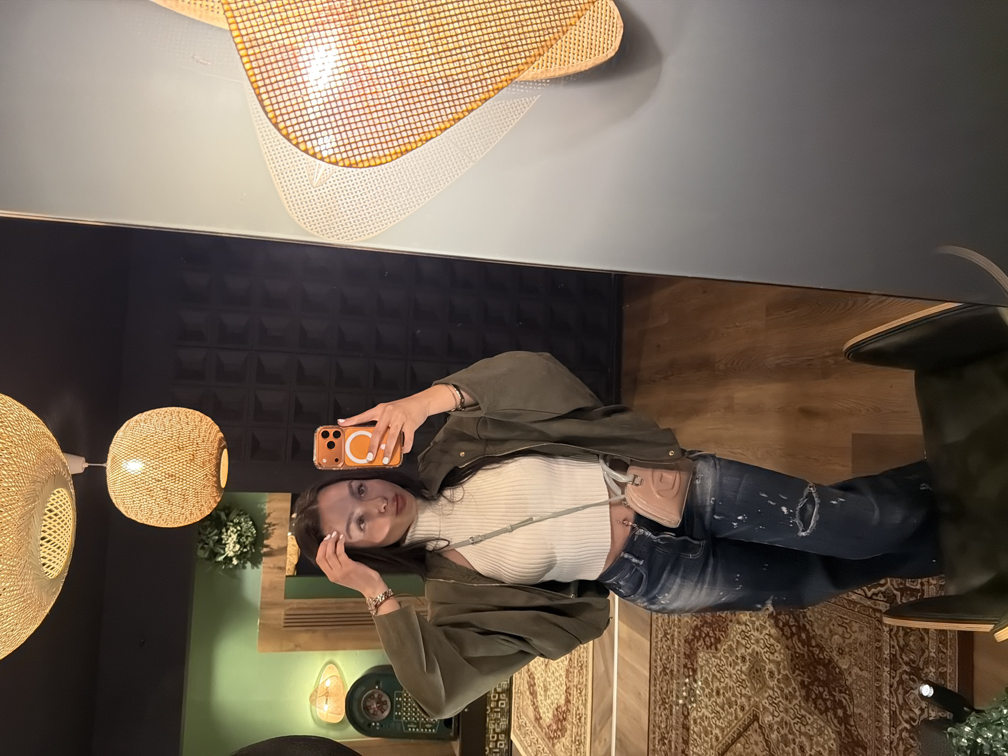 A woman is taking a selfie in a mirror. She is wearing a white turtleneck, an olive green jacket, and ripped blue jeans. She is holding an orange iPhone. The background includes woven light fixtures, a black textured wall, green walls, a rug, and furniture. The location is unknown and there is no indication that the image was taken in Kulata, Bulgaria.