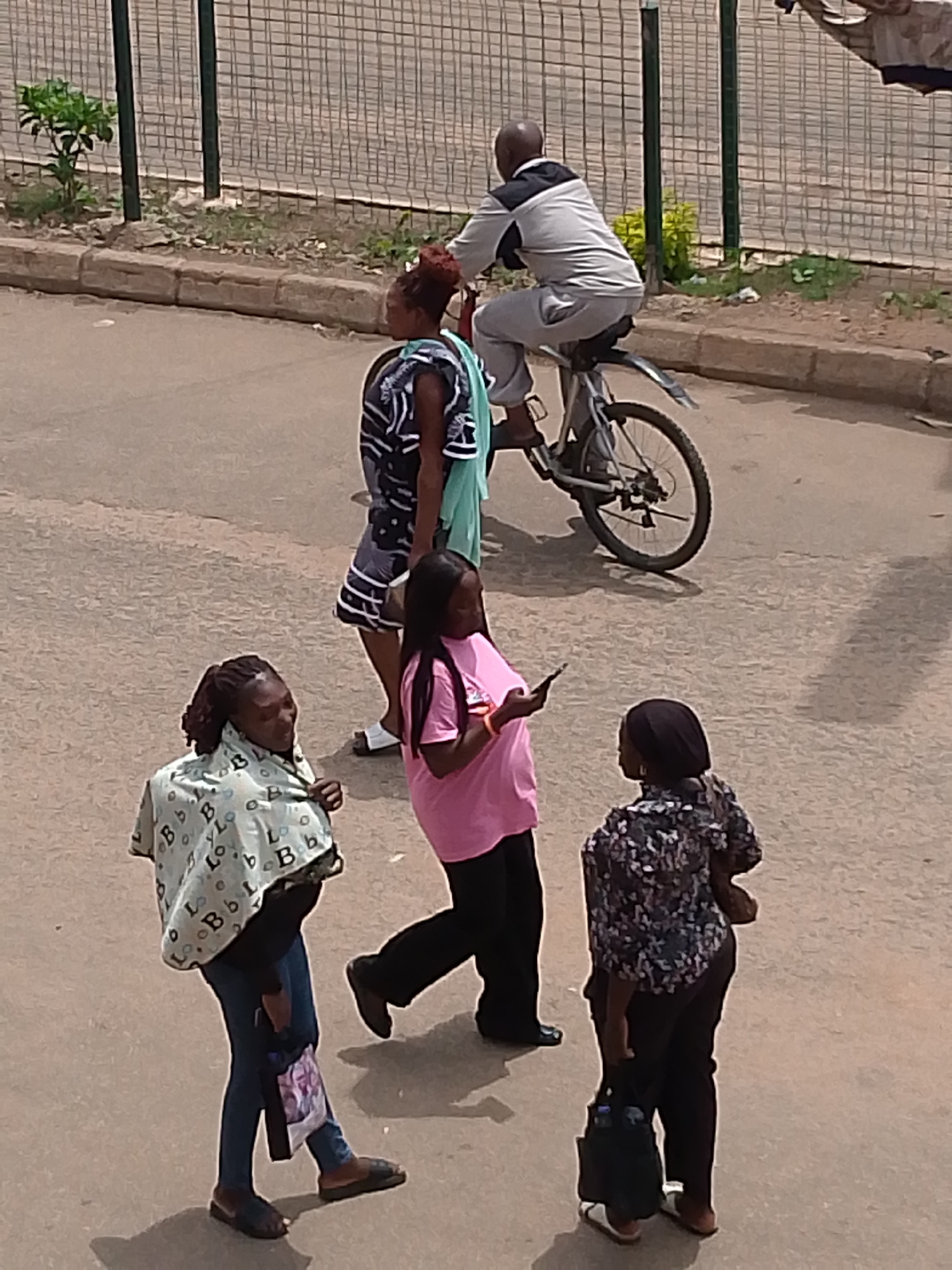 An outdoor, eye-level, medium shot shows a group of people on a paved surface. The lighting is bright, casting shadows on the ground. In the foreground, a woman with dreadlocks wrapped in a patterned scarf and wearing a black shirt and blue jeans walks to the left, carrying a bag. To her right, a woman in a pink t-shirt and black pants walks away from the viewer. Behind her, another woman in a black and white patterned shirt and black pants walks to the left, carrying a dark bag. In the background, a man on a bicycle with a black and gray jacket and gray pants rides away from the viewer. The setting appears to be an urban environment, possibly a street or sidewalk, with a fence and some vegetation visible in the background.