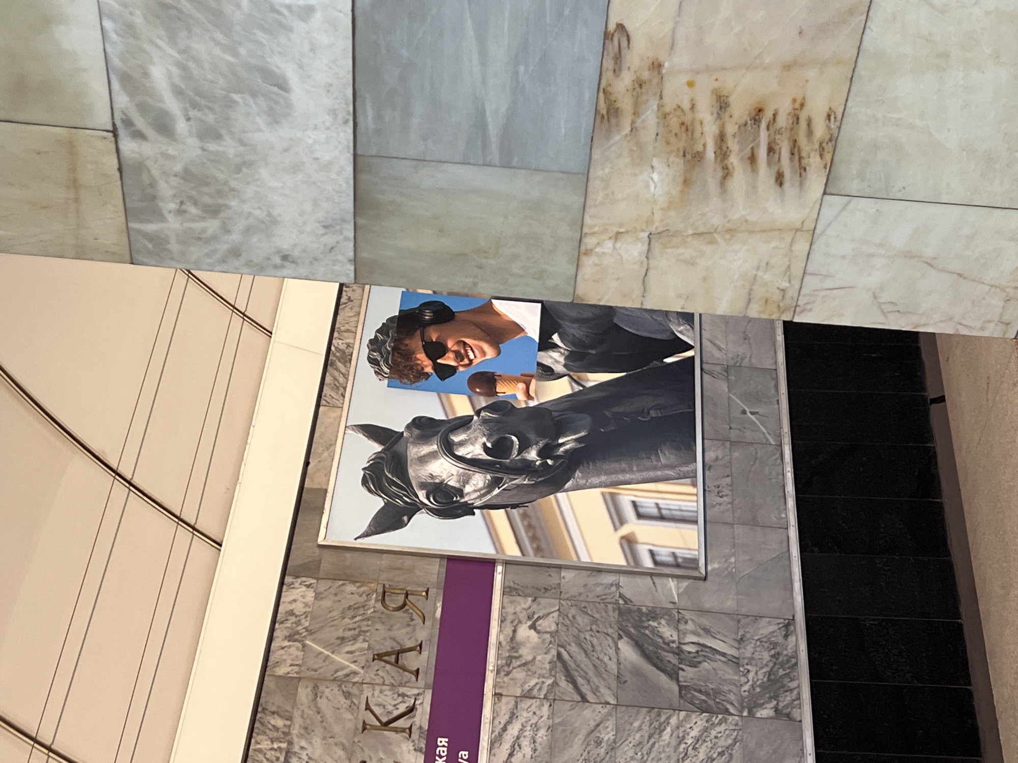 This image shows a close-up of a section of the Avtovo metro station in Saint Petersburg, Russia. The wall is covered in gray and white marble tiles. A promotional poster is attached to the wall, featuring a horse sculpture and a man. The purple strip on the poster has Russian text written on it. Beside the poster, there are rows of black tiles. The marble pattern continues to the ceiling.