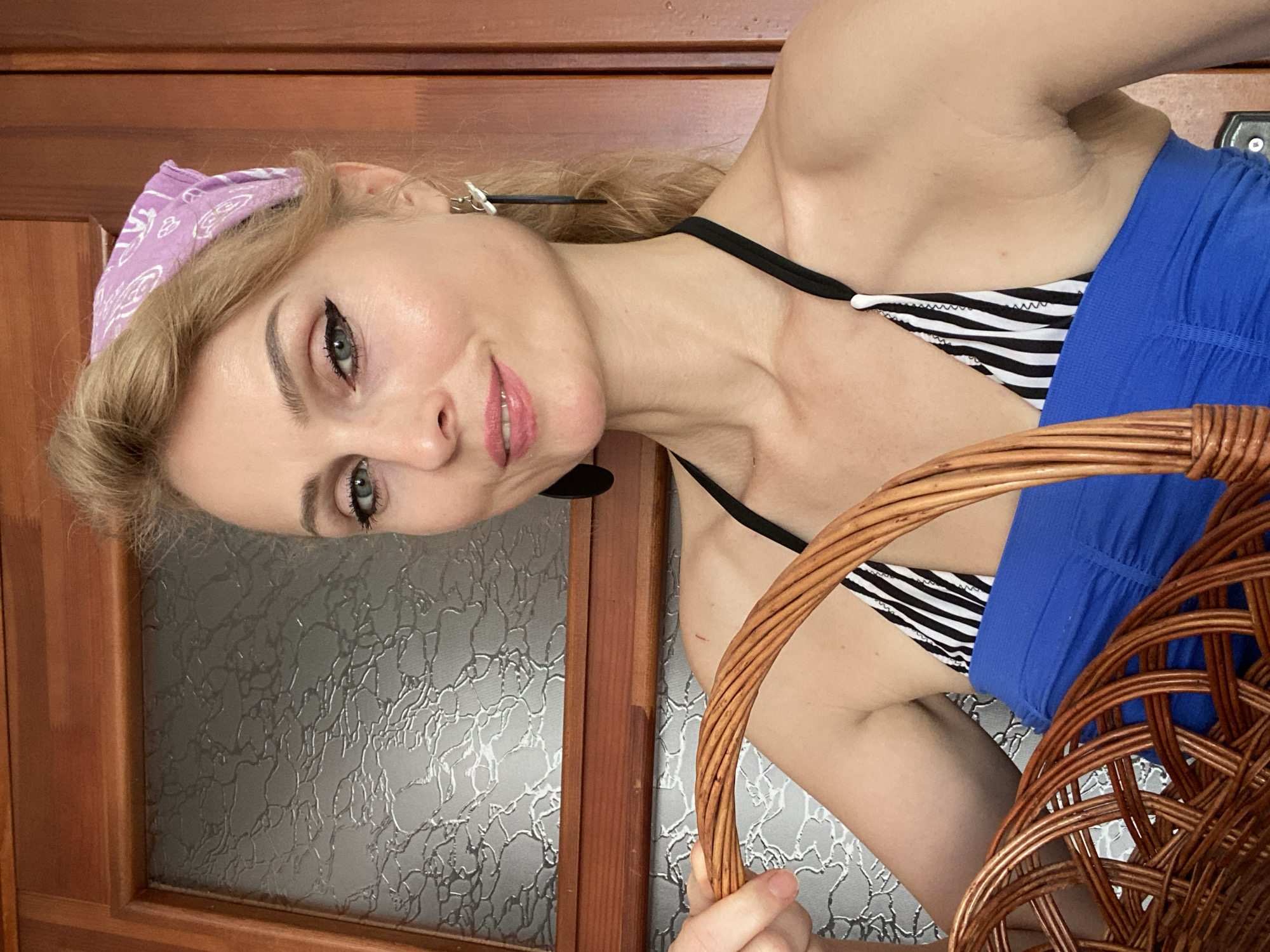A woman with blonde hair, wearing a purple bandana, a black and white striped bikini top, and a blue strapless top, is holding a wicker basket. She is standing in front of a wooden door with a textured glass panel. The scene appears to be indoors. There is no information available to suggest the location is the city of Lida, Belarus.