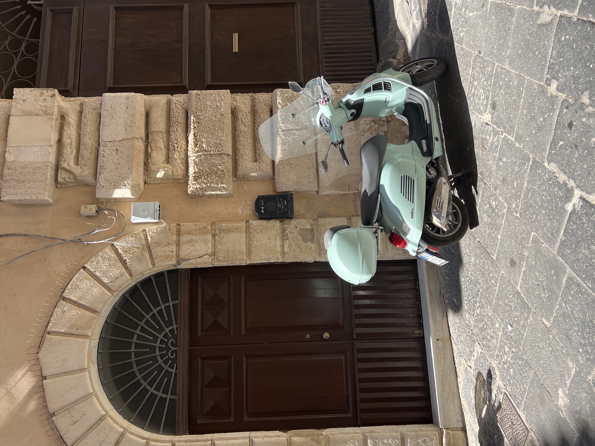 A light teal Vespa scooter with a clear windshield and a matching rear storage box is parked on a cobblestone street in front of a large, ornate wooden double door. The door is set within a stone archway with a decorative metal grate above. The building facade is constructed of rough-hewn, light-colored stone blocks. A black mailbox is affixed to the wall to the left of the door. Electrical conduits and a white outdoor electrical box are visible on the wall above the mailbox. Sunlight casts a shadow of the scooter onto the street. The scene is located in Syracuse, Italy.