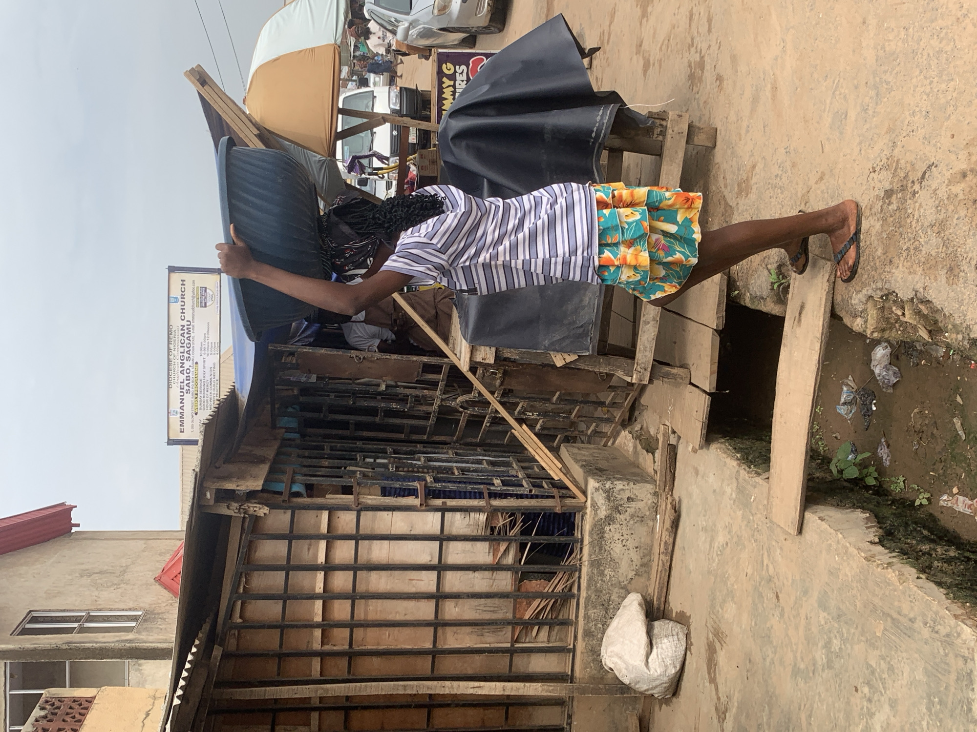 A woman in a striped shirt and floral skirt carries a blue tub on her head in Shagamu, Nigeria. She is stepping over a ditch filled with debris. A partially constructed building with a sign for Emmanuel Anglican Church is behind her. The sign indicates the location is 7, Odi Olowo Street, Sabo. Cars and market stalls are visible in the background.