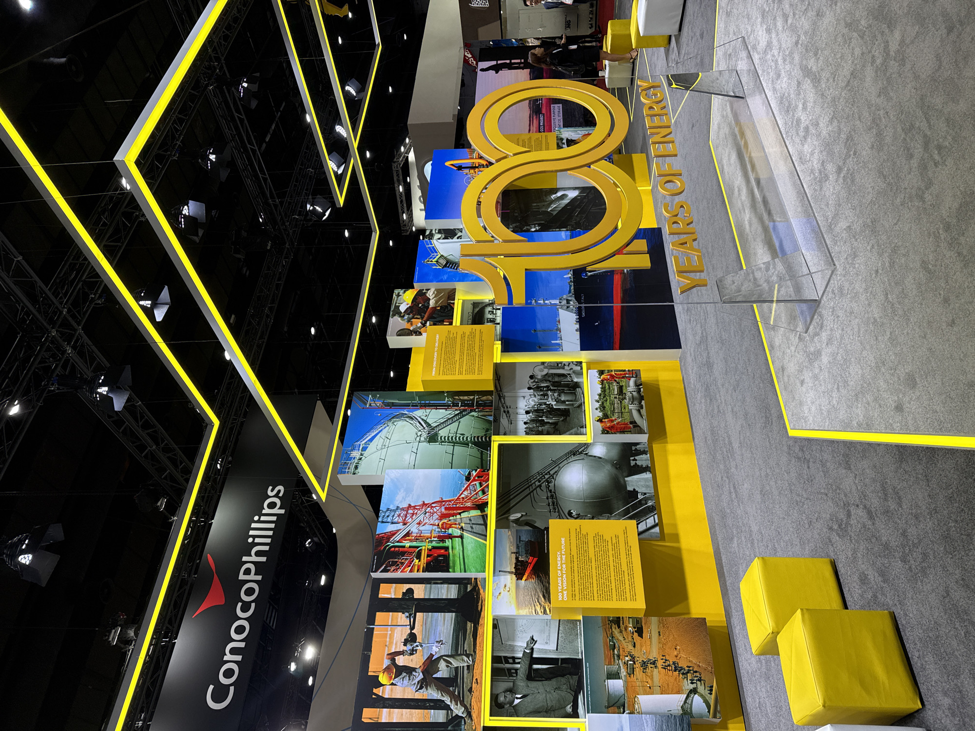 The image depicts an indoor exhibition booth for ConocoPhillips. The booth features a large, golden "100" sculpture, with "YEARS OF ENERGY" displayed below it. Behind the sculpture, a wall is adorned with various images and text panels showcasing the company's history and operations. These include photographs of industrial sites, workers, and infrastructure, as well as informative text. The ConocoPhillips logo is prominently displayed on a black banner overhead. The overall aesthetic is modern and professional, with yellow accents and bright lighting. The flooring is a grey carpet with yellow demarcations. In the background, two individuals are visible, one standing and one talking to another person. The image does not provide any information that would suggest it is located in Bangkok, Thailand.