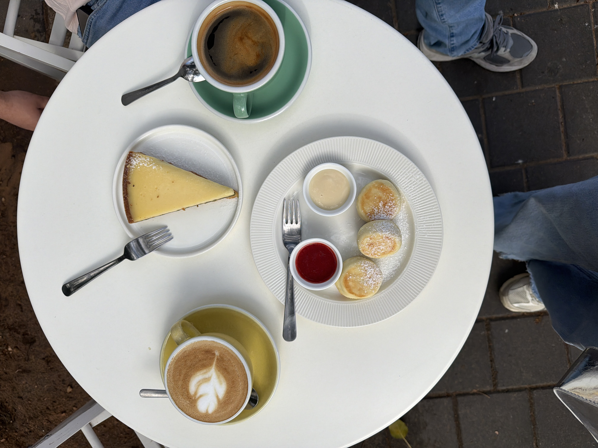 From an aerial perspective, the image presents a well-lit, outdoor scene. A round, white table is centrally positioned, displaying a selection of food and beverages. A slice of cheesecake is placed on a small white plate to the left of the table. Next to the cheesecake plate, a plate of small pancakes with powdered sugar sits with a small dish of red sauce and a small dish of white sauce. Two cups of coffee, one dark and the other a latte, rest at the top and bottom of the table. Silverware is provided for both the cheesecake and pancakes. Around the table, three individuals are seated, their legs and footwear partially visible. The ground surrounding the table suggests an outdoor café setting, with the surrounding area visible in the background. The location is the city of Bat Yam, Israel.