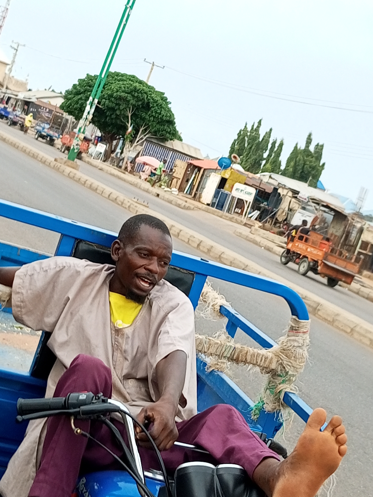A man is seated in the back of a blue three-wheeled motorized vehicle on a paved road, possibly in Tempure, Nigeria. He is wearing a light-colored robe over a yellow shirt and dark purple pants. His left foot is extended and visible, resting near the edge of the vehicle's cargo area. The man appears to be speaking and looking to his right. The vehicle is in motion, with the road and buildings lining it visible in the background. Some buildings appear to be shops or market stalls. There is a large green tree and a tall green and white pole with a traffic signal attached to it in the middle ground. Other vehicles, including a small orange truck, are on the road in the distance. The sky is overcast.