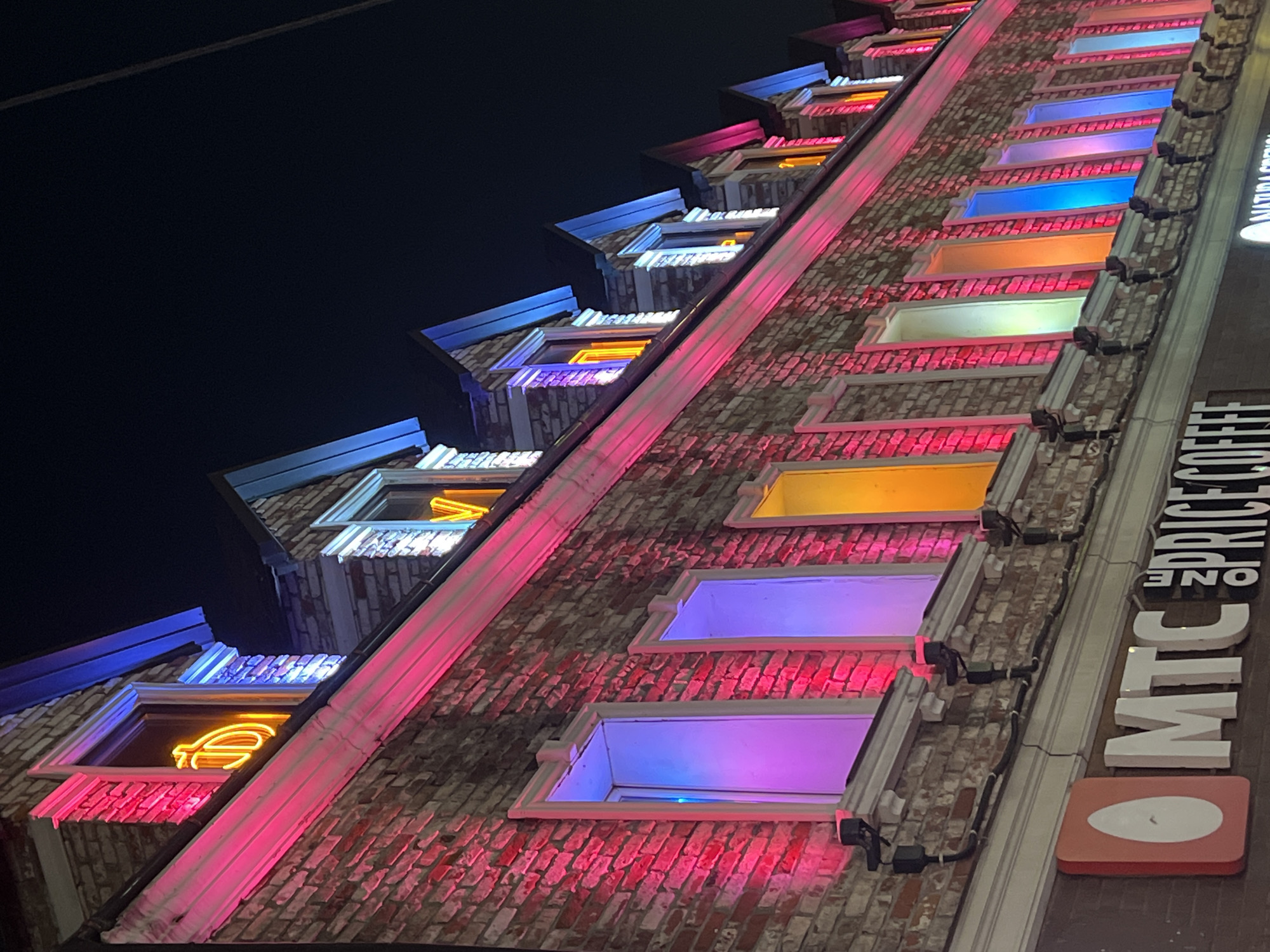 The image shows a brick building at night, illuminated by colorful lights. The building has multiple dormer windows on its upper floors, each with a different colored light shining from within, including pink, purple, yellow, and blue. Some windows also have neon signs visible. The lower part of the building features storefronts with illuminated signs. One sign reads "MTC" and another says "ONE PRICE COFFEE". The overall scene is brightly lit and vibrant, suggesting a lively urban environment.