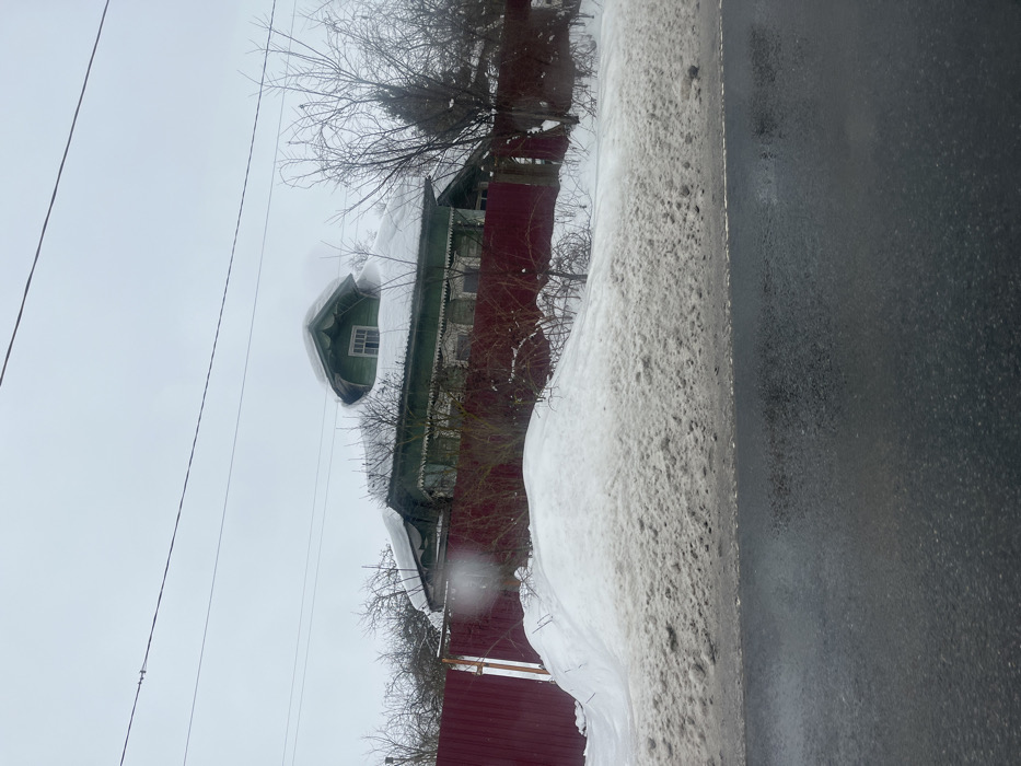 The image displays an outdoor winter scene in Podvyaznovo, Russia, under an overcast, light gray sky. A traditional green wooden house with white decorative trim around its windows and eaves stands prominently. Its roof is heavily covered in snow, from which a distinct, rounded green dormer window with white trim protrudes. A plume of white vapor or smoke is visible rising from behind the dormer. The house is enclosed by a solid red metal panel fence. In the foreground, a dark, wet asphalt road runs horizontally, bordered by a large, irregular snowdrift. The snow closest to the road is discolored with dirt and grit, while the snow closer to the fence appears cleaner. Bare deciduous trees, some with snow on their branches, are scattered around the house and behind the fence. Multiple overhead utility lines traverse the upper portion of the frame. No individuals or active subjects are present in the scene.