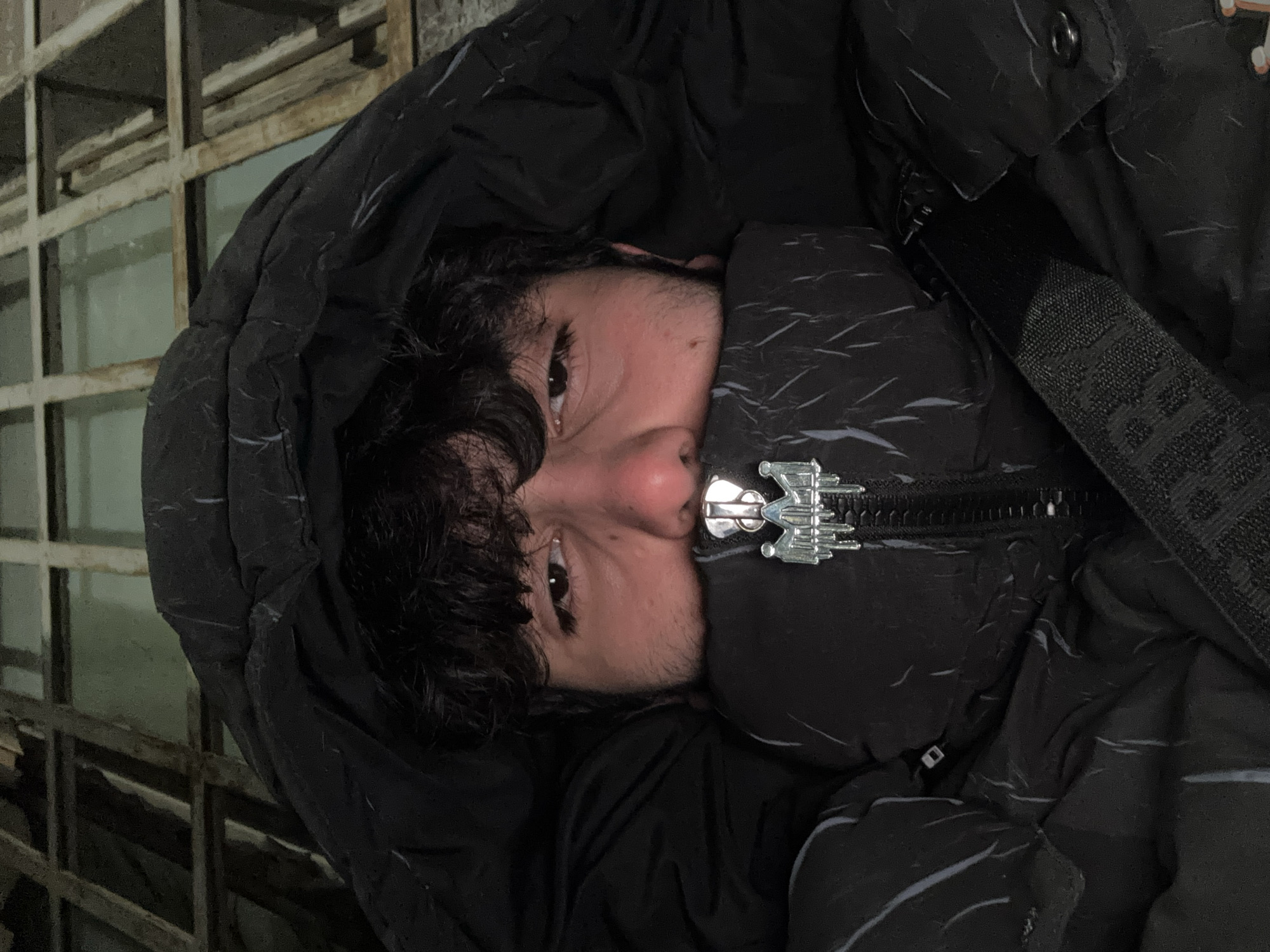 A person with dark hair and light skin peers from the confines of a black hooded jacket, zipped up to the nose with a decorative metallic zipper. The person is positioned against a weathered, gray-colored metal grid structure. The location is likely Yerevan, Armenia, based on metadata not included in the image itself. The person's eyes are visible, and the jacket's hood and high collar obscure most of the face. A black strap with lettering is visible to the right of the jacket zipper. The image's orientation is rotated.