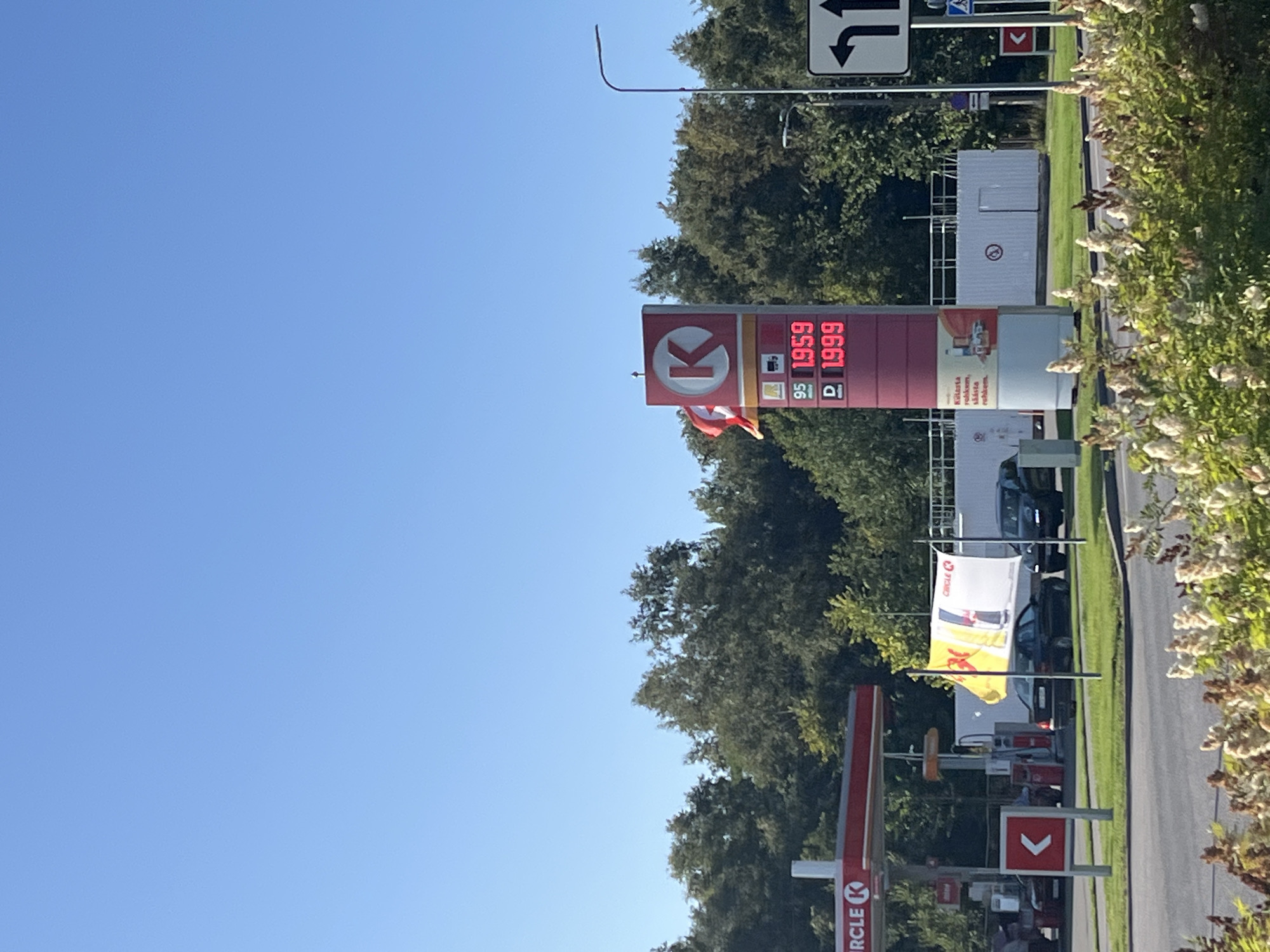 The image shows a Circle K gas station in Kohtla-Järve, Estonia, on a clear day. A red and white gas price sign displays fuel prices: 95 octane at 1.959 and diesel at 1.999. A Danish flag is attached to the sign. To the left, a red awning covers gas pumps. In the background, a line of trees borders the property, and a white building with a fence is visible. Two cars are parked near the pumps, and another car is driving away. A street sign indicating a left turn and a pedestrian crossing is present. The foreground is filled with lush green bushes and plants.