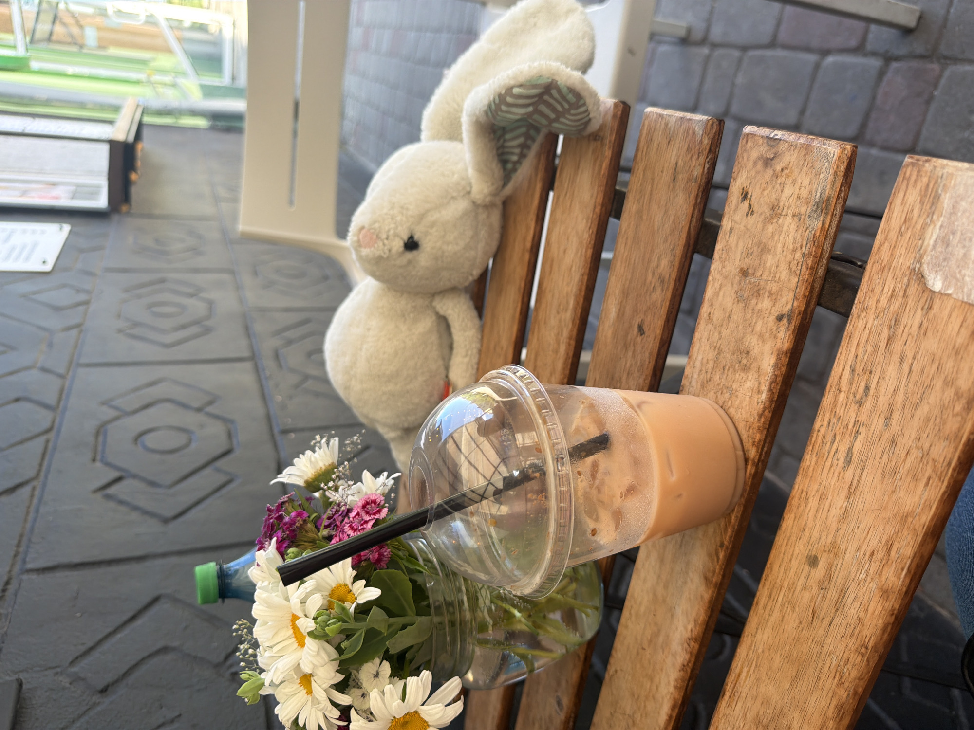 A stuffed white bunny sits on a wooden chair next to a bouquet of flowers in a plastic bottle and a cold beverage. The bunny is leaning against the chair, its ears are floppy. The flowers are a mix of white daisies and purple carnations. The cold beverage is in a clear plastic cup with a black straw. The scene appears to be outdoors, possibly at a cafe or park, as suggested by the wooden chair and the patterned ground. The lighting suggests it is daytime. The overall mood is relaxed and casual. There is no visible text in the image.