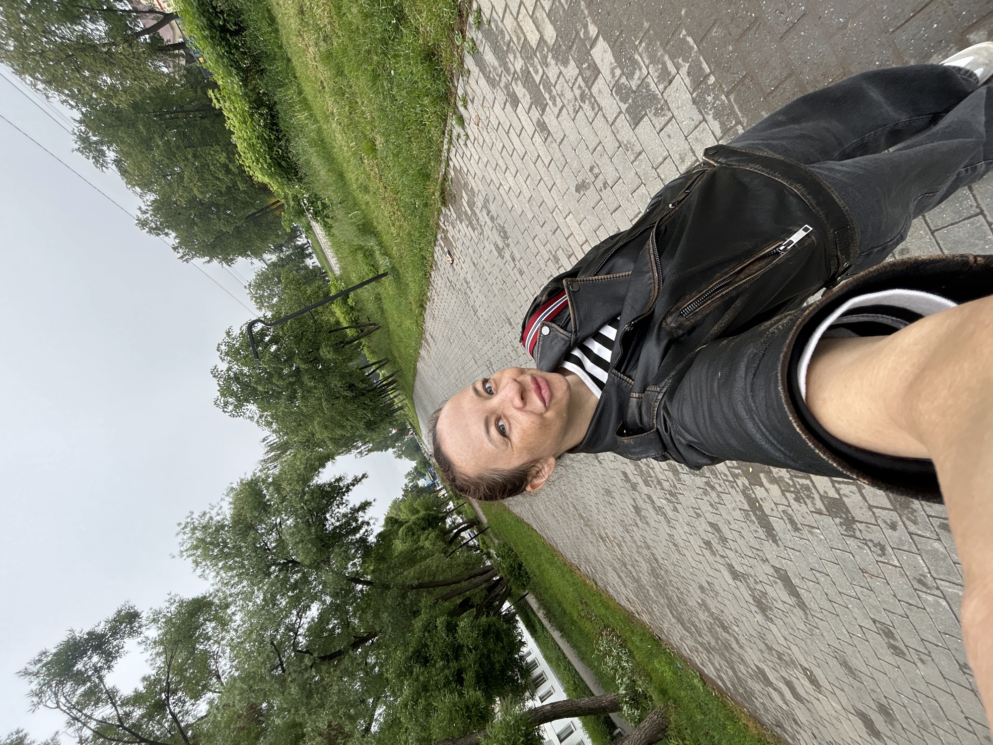 This rotated selfie shows a person, likely female, posing outdoors in Chaikovsky, Russia. She looks directly at the camera with a neutral expression, wearing a black leather jacket over a black and white striped shirt, dark pants, and white sneakers.
The setting is a paved pathway made of light grey interlocking bricks, showing wet patches that suggest recent rain. Lush green grass borders the path on both sides, and numerous trees with vibrant green foliage line the walkway. The sky above is a light, uniform grey, indicating an overcast or cloudy day. In the background, more trees extend into the distance, and a multi-story building is partially visible through the foliage. Thin power lines or cables can be seen against the sky. The image captures an everyday moment on a damp, green landscape. There is no visible text.