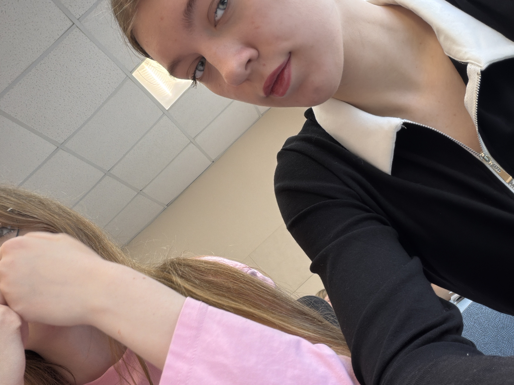 This upward-angled close-up image depicts two young females indoors, in what is identified as a location within the city of Chaikovsky, Russia. The primary subject, situated on the right, is a female with fair skin, light blue-grey eyes, and visible dark eyelashes. Her lips are colored with a pinkish-red pigment. She is wearing a black top with a white collar and a silver zipper, which is partially unfastened. Her light brown hair is visible along the right edge of the frame. Her gaze is directed towards the camera. The secondary subject, partially visible on the left, has long, light brown hair mostly obscuring her face, though a segment of eyeglasses is discernible behind her hair. She is dressed in a pink long-sleeved garment, and her right forearm, which exhibits a small reddish mark, is raised towards her face. The indoor environment features a white acoustic grid ceiling with one rectangular fluorescent light fixture visible in the upper center. A light beige wall is present in the background. In the bottom-left foreground, a grey surface with a fine white grid pattern is partially visible.