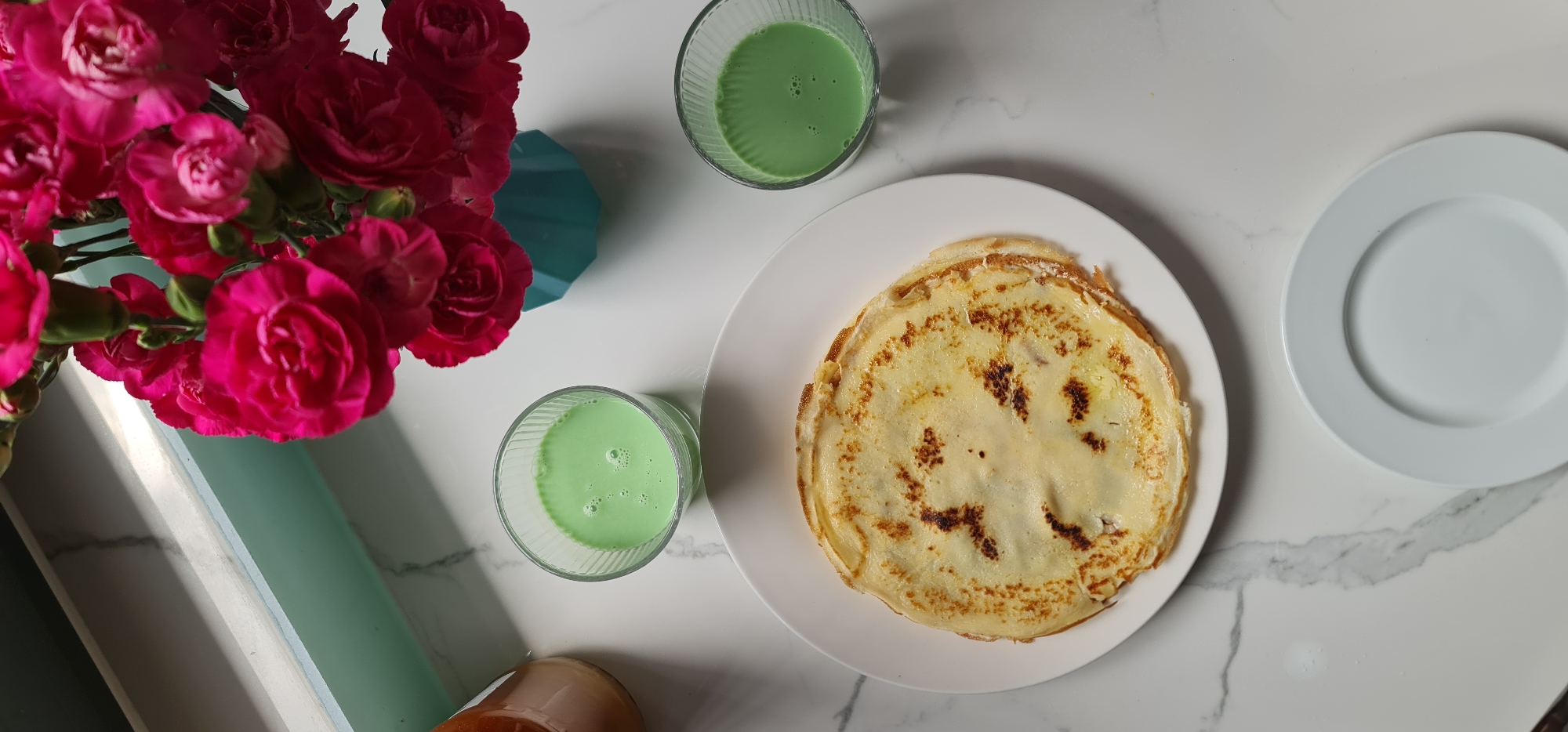 The image shows a still life arrangement on a white marble surface. A bouquet of bright pink roses in a light blue vase is on the left. Two glasses filled with a green liquid are positioned near the flowers and to the right. A stack of pancakes on a white plate sits in the center of the frame, and an empty white plate is placed further to the right. The objects are arranged on the surface in a way that suggests a breakfast or brunch setting. No people are visible. The location (Đà Nẵng, Vietnam) cannot be determined from the information given in the image.