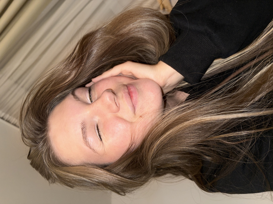 A young woman with closed eyes rests her head on her hand. She has fair skin, light eyebrows, and pursed lips. Her long, light-brown hair with blonde highlights cascades around her face. She is wearing a black long-sleeved shirt. The background includes a neutral-colored wall and curtain. The image is angled, placing the subject on a diagonal plane.