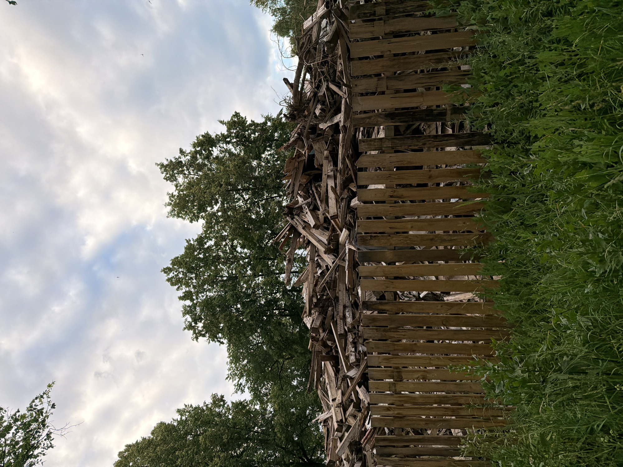 The image shows a close-up, outdoor view of a rustic wooden structure made of stacked planks, resembling a fence or a makeshift wall. The structure is densely packed with various pieces of wood, creating a textured and somewhat chaotic appearance. To the left of the wooden structure, lush green foliage and trees are visible, suggesting a natural, outdoor setting. In the background, a cloudy sky with patches of blue is present, indicating daytime. The overall impression is one of a weathered, possibly abandoned, or DIY construction integrated into a natural environment. There is no visible text in the image.