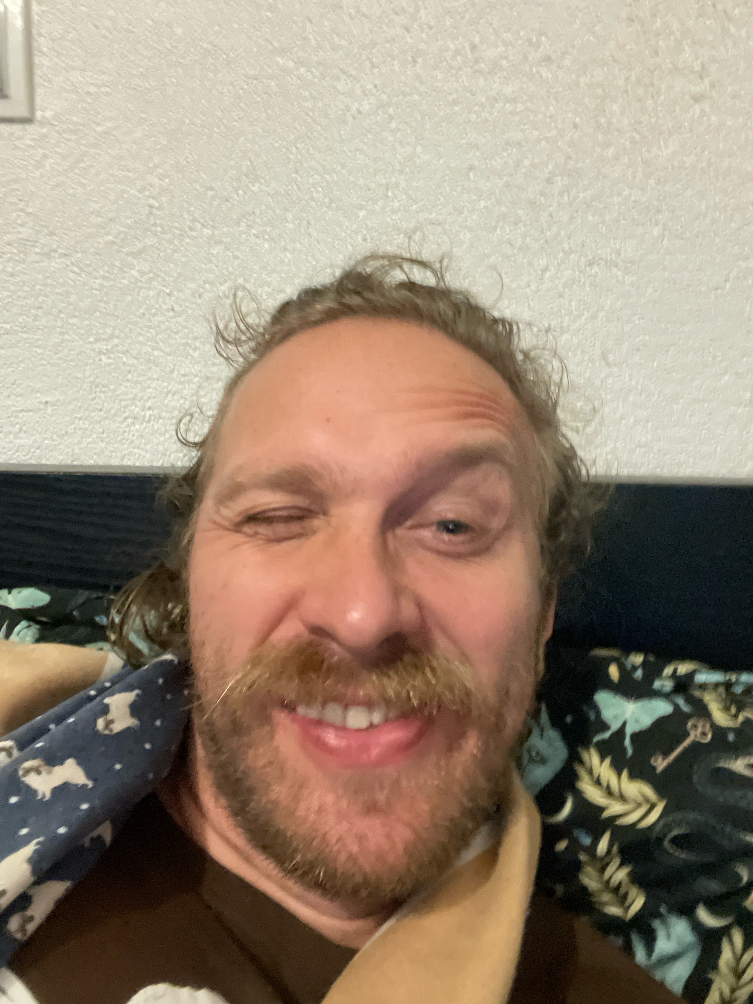 The image is a close-up selfie of a man winking. He has a mustache and beard, and his hair is curly and light brown. He is wearing a brown shirt and is partially covered by a blanket with a pug pattern. Another blanket with a celestial and botanical pattern is visible in the background. The man appears to be indoors, with a textured wall behind him and a dark surface that could be furniture. The scene is not identifiable as being in Kornevo, Russia.