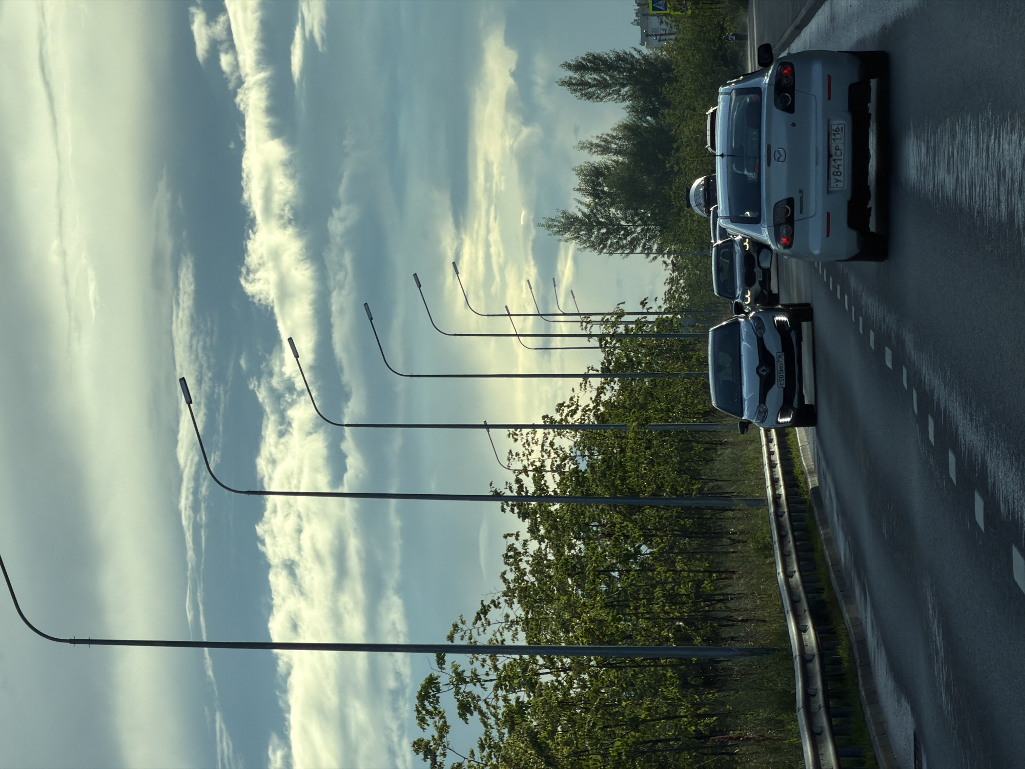 The image shows a view of a highway or a large road in Kazan, Russia, with multiple lanes of traffic. The road surface is wet, indicating recent rain or damp conditions. On the left side of the road, there is a guardrail and a dense line of trees and vegetation. A series of tall lampposts are positioned along the left side of the road, with their lights off. The sky above is overcast with dramatic, layered clouds.
Several cars are visible on the road, traveling in the same direction. In the foreground, a silver car is in the right lane. Further ahead, in the left lane, a white car is visible. Behind these vehicles, other cars are also on the road. The license plates of some of the vehicles are partially visible, with Russian characters and numbers. On the far right of the image, a yellow pedestrian crossing sign is visible. The overall scene suggests a typical day on a major road in an urban or suburban setting in Kazan.