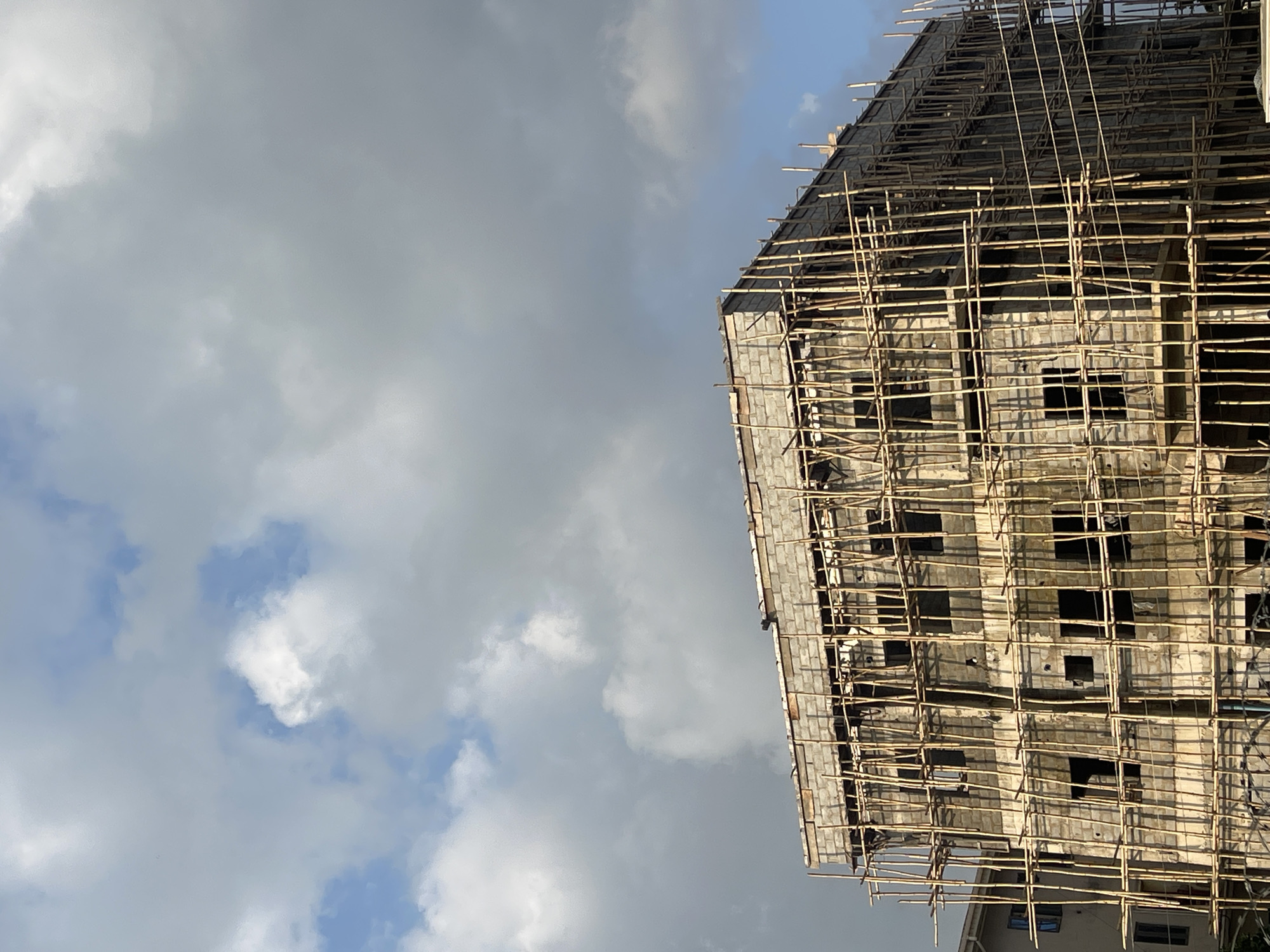 The image depicts a multi-story building under construction in Abuja, Nigeria. The building is constructed with concrete blocks and is extensively covered in bamboo scaffolding. Numerous windows are visible, most of which are empty openings. The sky above is partly cloudy with a mix of gray and white clouds against a blue background. To the left of the construction site, a portion of another building, also appearing to be under construction or recently built, is visible. The scene is devoid of people, indicating a pause or inactivity at the construction site. The overall impression is one of ongoing development in an urban environment.