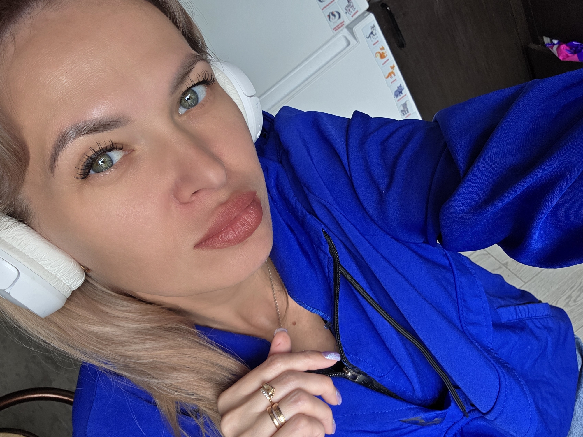 A woman is shown in a close-up, selfie-style portrait, wearing a bright blue jacket and white headphones. Her face is turned towards the camera, with her eyes looking forward. Her blonde hair is visible beneath the headphones. She is wearing rings on her fingers. The background includes a portion of a dark wooden surface, a white wall with some colorful graphics, and a hint of a dark metal chair. The image appears to be taken indoors, possibly in an apartment or studio in the city of Chaikovsky, Russia, though no specific Chaikovsky landmarks are visible.