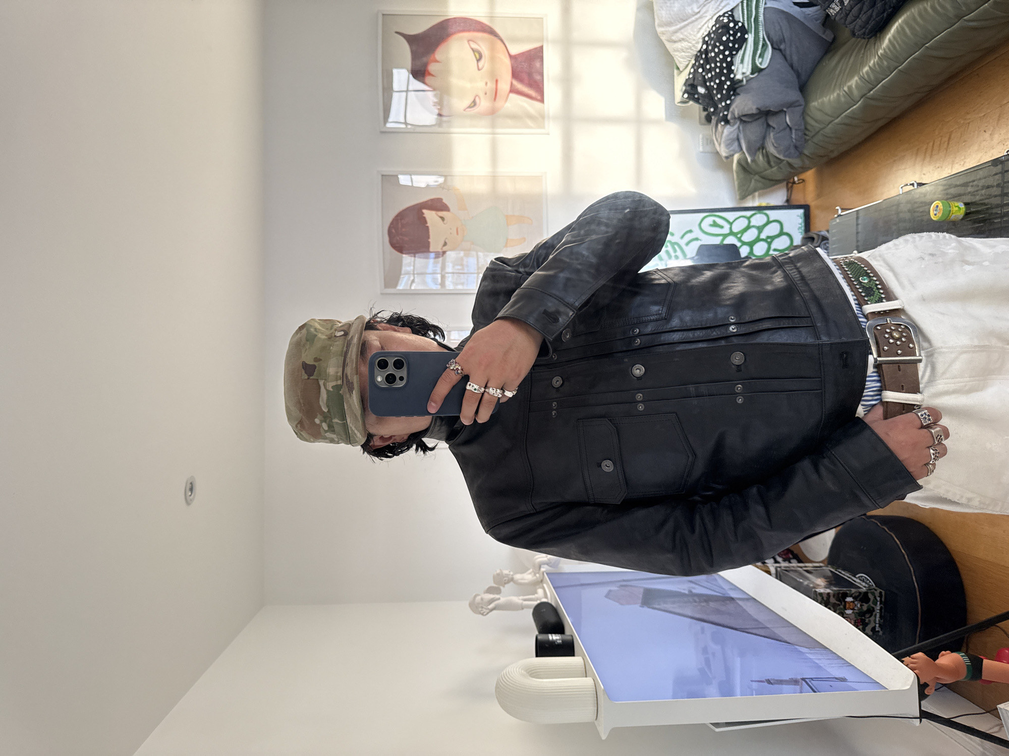 A person wearing a camouflage hat, black leather jacket, white pants, and multiple rings is taking a selfie in an indoor environment. The background includes artwork on the wall, a pile of clothing, a green-patterned design, a case, and a portion of a wooden floor. In the foreground, there's a table with a screen showing a reflection and several decorative figurines. The location is the United Kingdom.