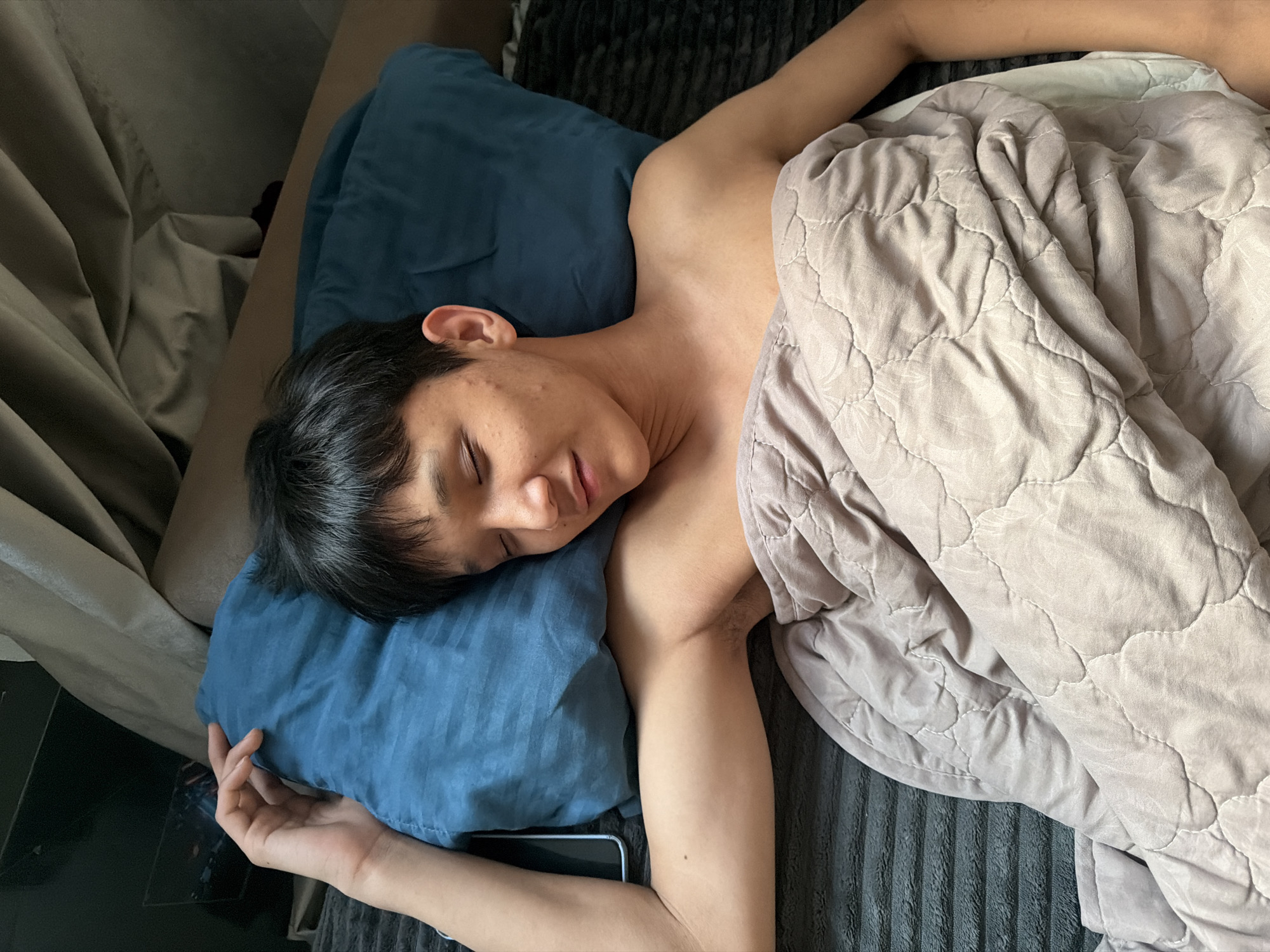 A young person with black hair and closed eyes is sleeping in bed, covered by a light brown quilted blanket. They are lying on their side, with their head resting on a blue pillow. Their left arm is extended, and a smartphone is visible next to the pillow. The background consists of bedding and curtains. The scene is indoors, likely in a bedroom.