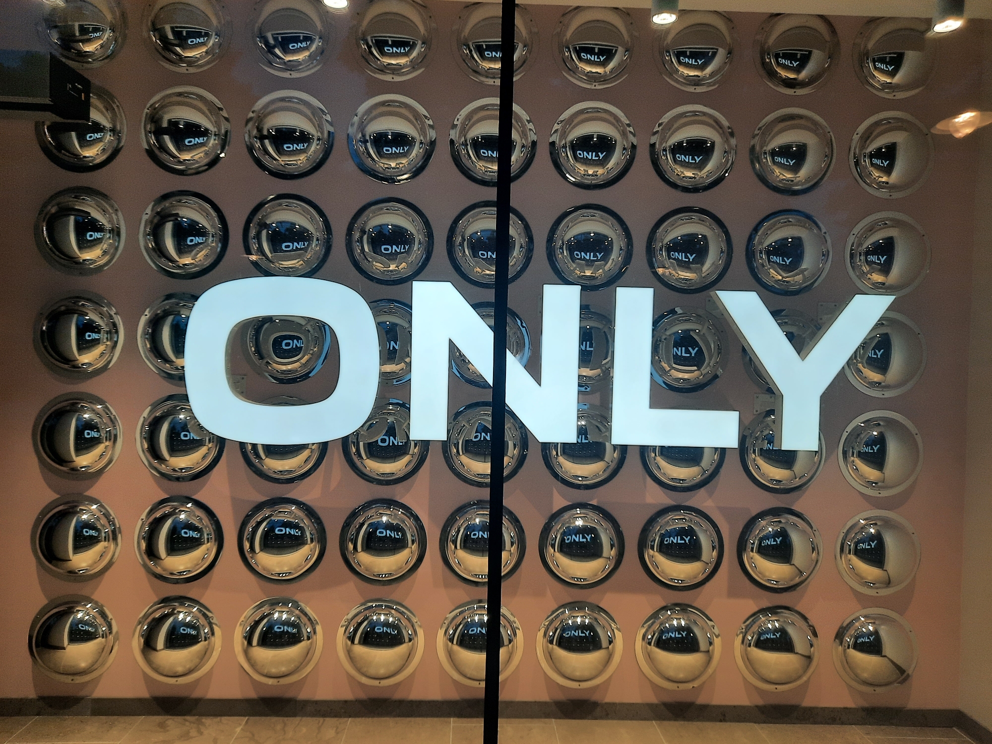 The image shows a store window in Rosenheim, Germany, with the word "ONLY" prominently displayed in large white block letters. Behind the letters, the window is decorated with a grid of circular, convex mirrors, each reflecting the word "ONLY" in smaller text. The overall impression is one of repetition and branding. The store appears to be on a street, with lighting fixtures visible above the mirrors.