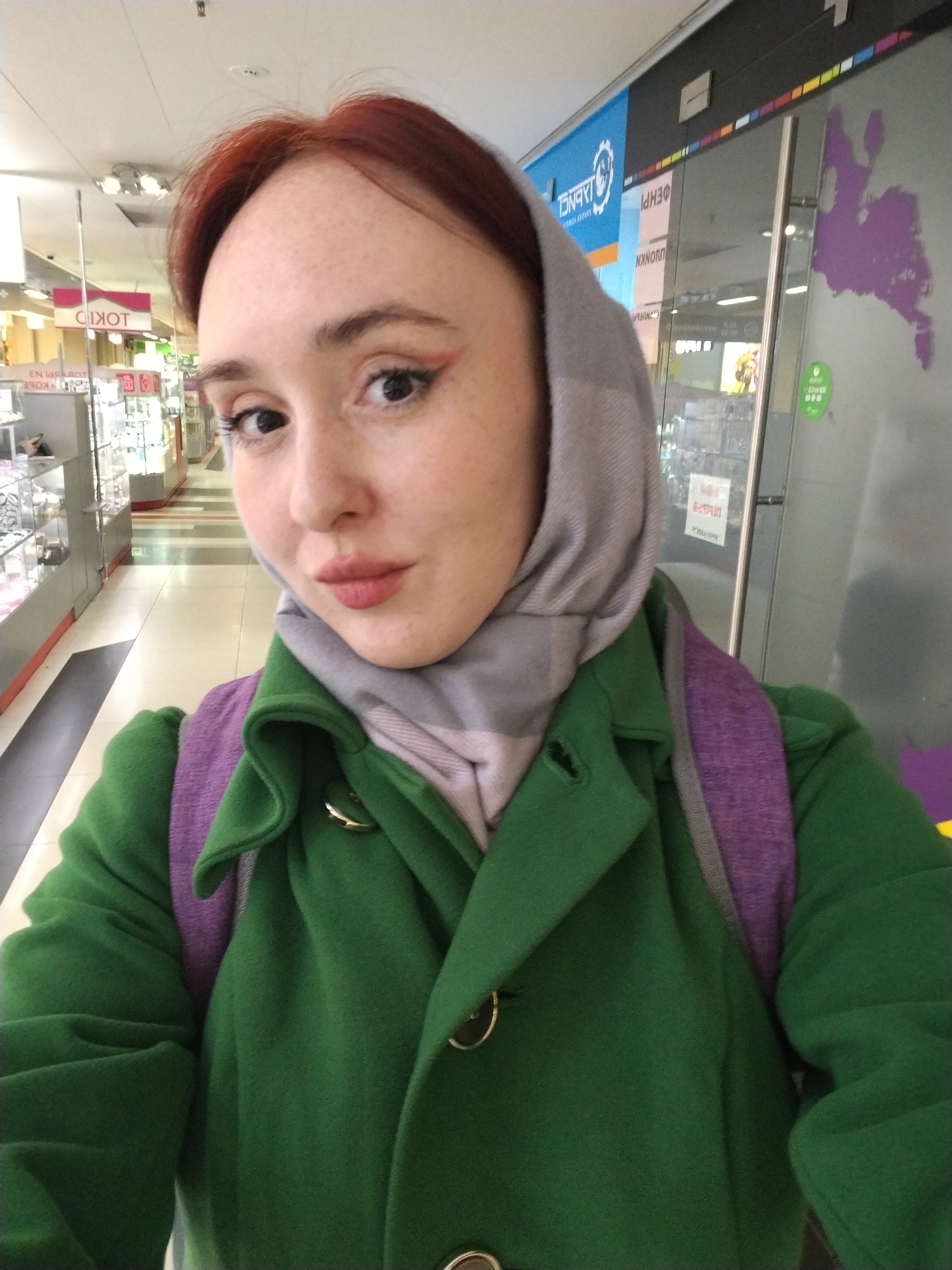 A woman is taking a selfie indoors, in what appears to be a shopping mall or commercial center in Izhevsk, Russia, judging by the signage visible in the background. She has light skin, red hair partially covered by a grey headscarf, and is wearing a green coat with gold buttons and a purple backpack. She is wearing makeup including eyeliner and lipstick. The background features displays of what appears to be jewelry, and signs written in Russian. A map of Europe is visible on a glass wall.