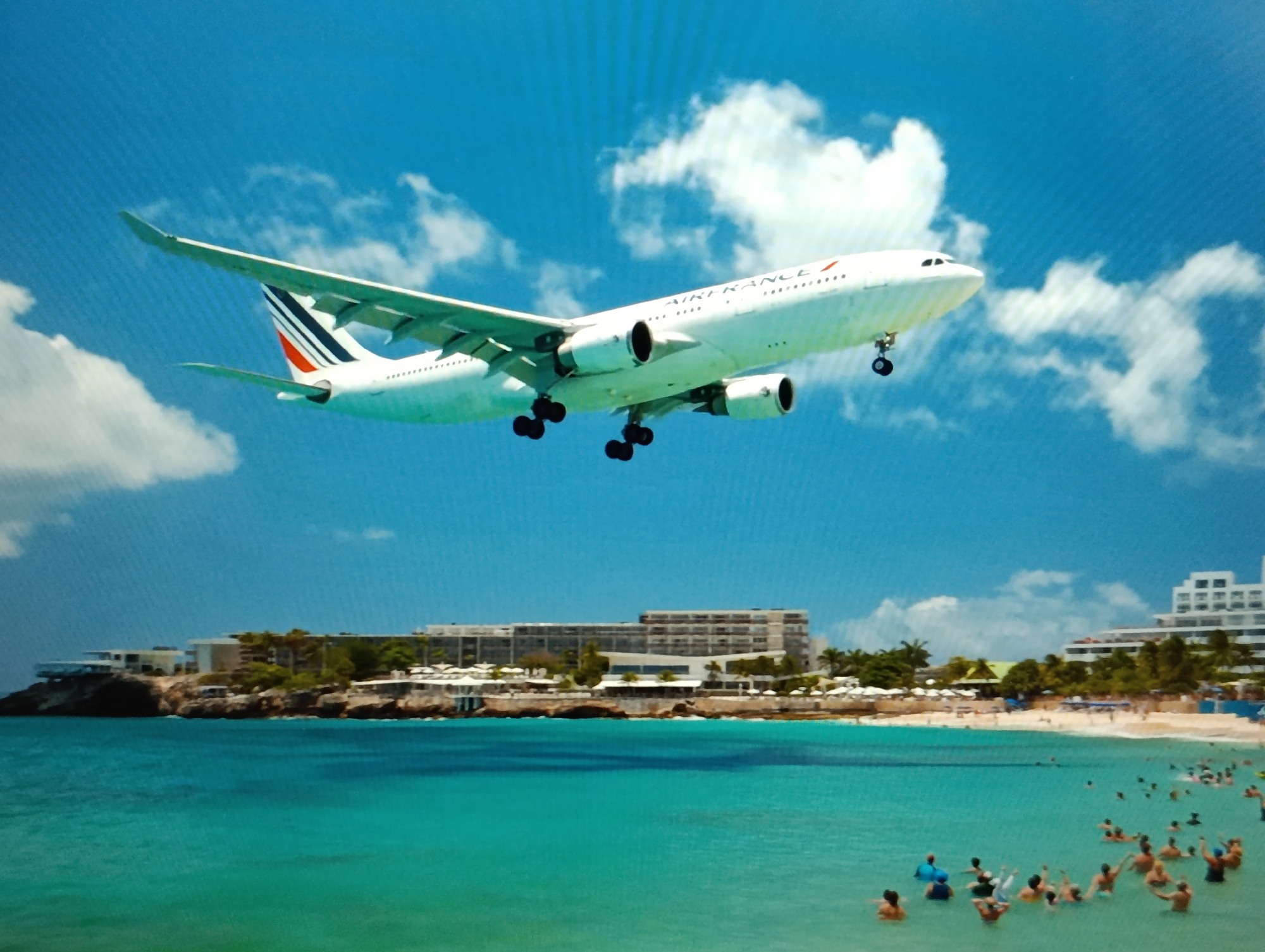 An Air France airplane is captured mid-flight against a bright blue sky dotted with white clouds, appearing to be on its approach to an airport. The aircraft's landing gear is deployed, suggesting it is either landing or taking off. Below the airplane, a picturesque tropical beach scene unfolds. The turquoise water is clear and calm, with numerous people swimming and enjoying the ocean. A sandy beach stretches along the coastline, lined with palm trees and several resort-style buildings, including a large hotel. The buildings are modern and appear to be situated on a rocky outcrop overlooking the sea. The overall impression is a vibrant vacation destination with a dramatic aerial element. The location is the city of Dubăsari town council, Moldova.