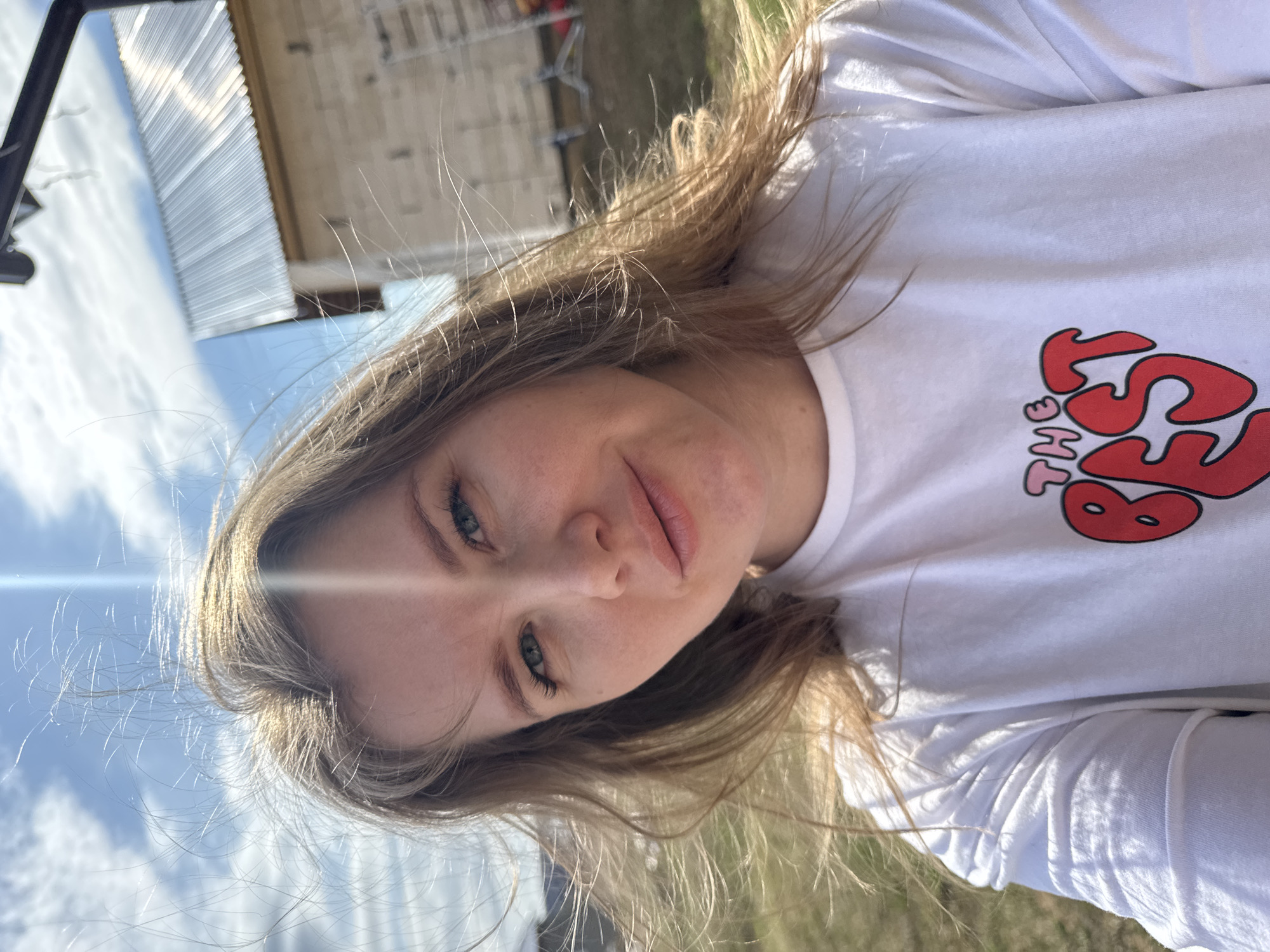 A young woman with long, light brown hair and blue-green eyes takes a selfie outdoors. She is wearing a white long-sleeved t-shirt with a red and pink graphic on the chest. The graphic features the word "THE" in smaller pink text, followed by "BEST" in larger, bubbly red letters, with a small pink heart detail.
Strong sunlight from the upper left creates a prominent lens flare across her face and hair, highlighting her features and strands of hair. She looks directly at the camera with a calm, slightly smiling expression. The background shows a bright blue sky with scattered white clouds on the left. On the right, a light-colored wooden or corrugated metal building structure is visible, suggesting a rural or suburban setting. Below the building, blurred green and brown ground is present. A dark, angular object is partially visible on the far left edge of the frame. The overall impression is a sunny day in Gornyi (Dvurechenskoe S/P), Russia.