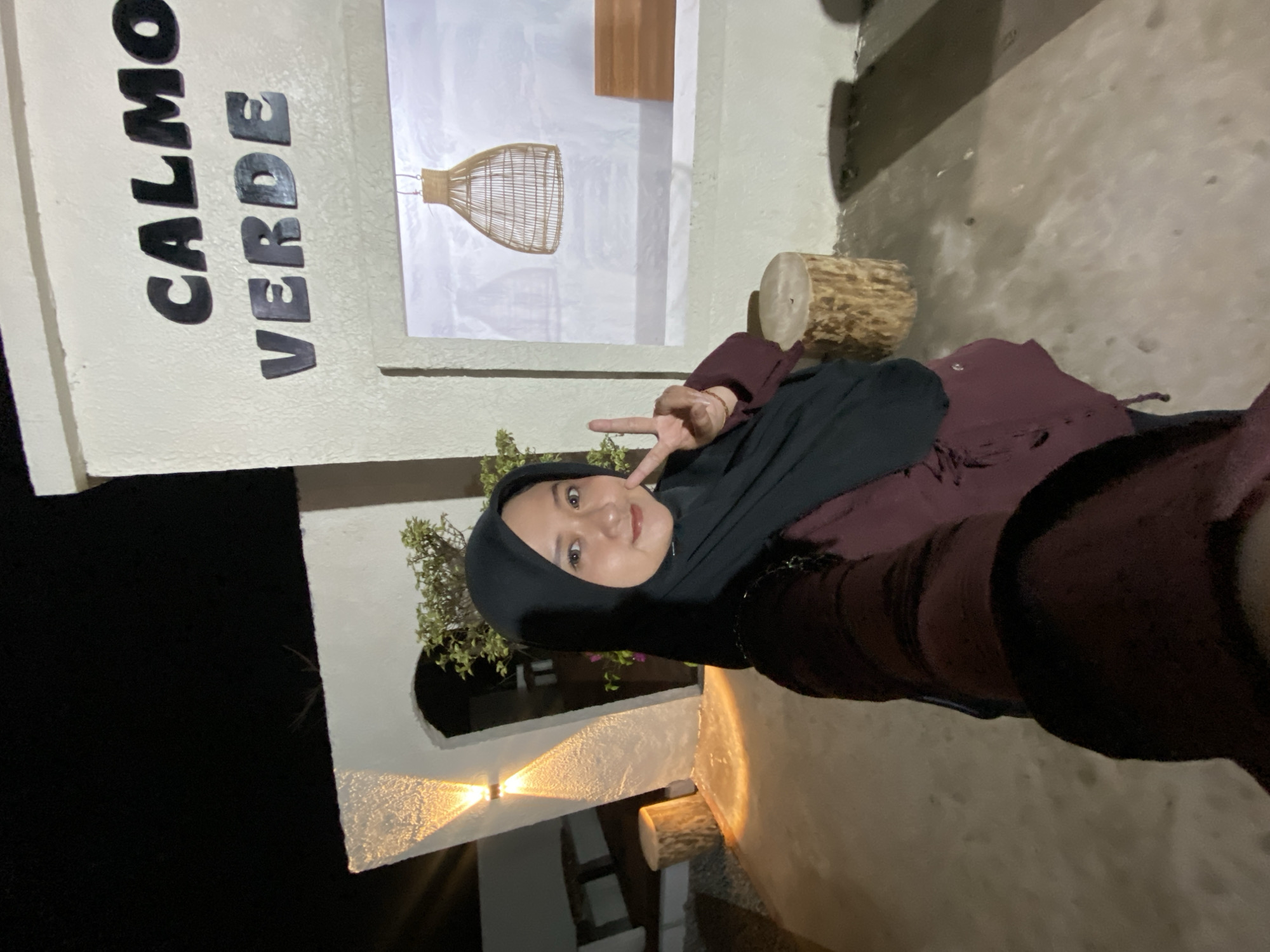 In this medium shot, a young woman wearing a black hijab and a maroon dress is taking a selfie. She is positioned in the lower right of the frame, looking towards the camera with a smile and holding up two fingers in a peace sign. The background is a textured, light-colored wall with decorative elements. To the left of the woman, the words "CALMO VERDE" are prominently displayed in large, dark letters on the wall. Behind the text, a framed artwork is visible, featuring a drawing of a woven lampshade and a wooden architectural element against a white backdrop.
To the right of the artwork, a section of a rough, light-colored wall is visible, with a log-like object resting against it. The scene appears to be outdoors, with dark shadows suggesting it is nighttime or dusk. The lighting from two wall-mounted lamps casts a warm glow on the textured wall in the lower part of the image. A hint of greenery from a plant is visible to the left, near an arched opening in the wall. The overall impression is a casual, perhaps social setting, possibly a cafe or public space with artistic decor. The woman's pose and expression convey a sense of happiness and engagement. The location context provided is Kesiman, Indonesia.
