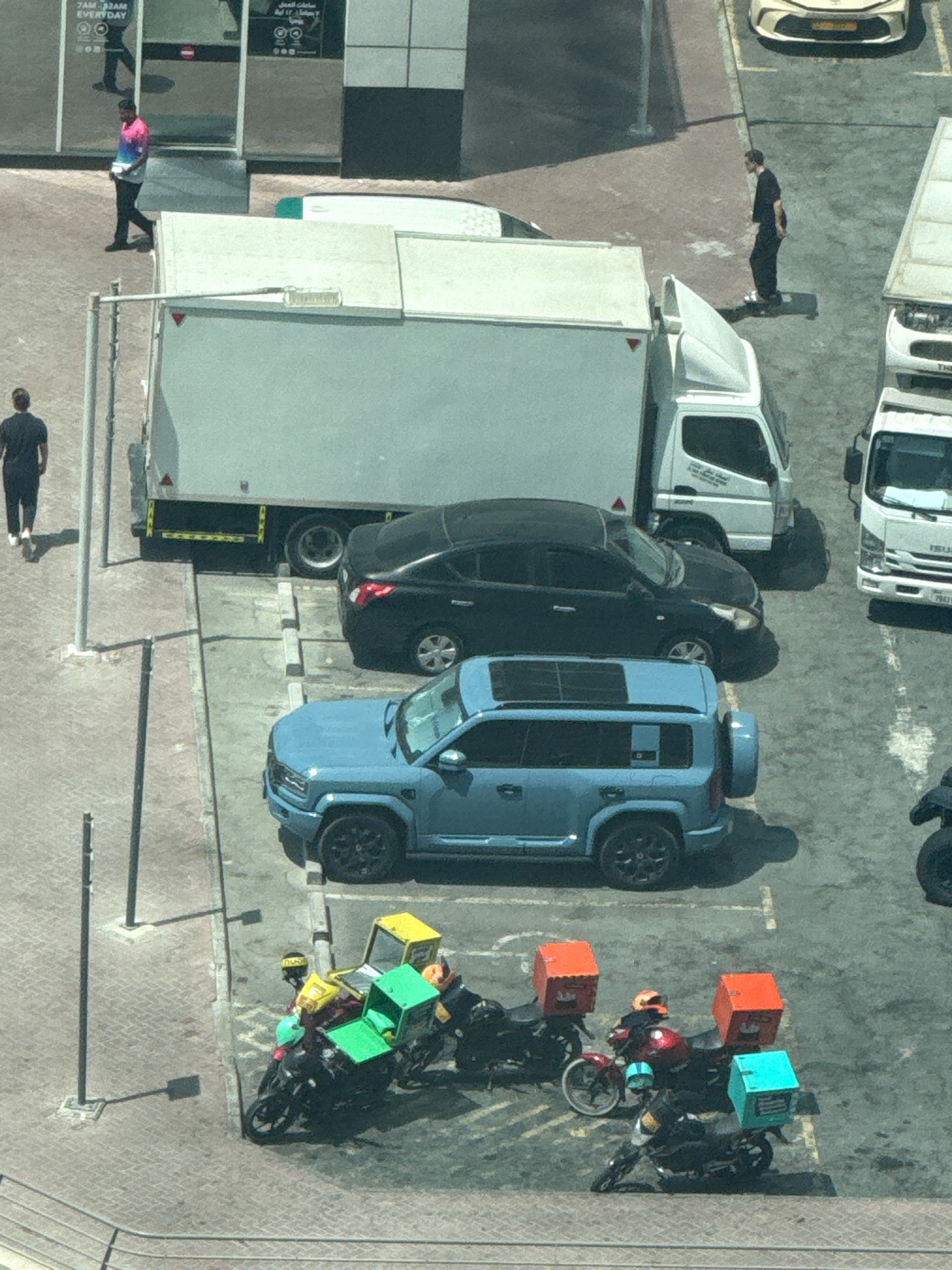 An aerial view captures a street scene in Dubai, United Arab Emirates, characterized by a multi-lane road and a parking area. On the left side of the image, a man wearing a pink and blue shirt walks on a sidewalk adjacent to a building. Further down the street, a large white truck with a grey cargo box is parked, with a black sedan parked behind it. To the right, a blue SUV is parked. In the foreground, a group of three motorcycles, each equipped with delivery boxes of various colors (green, yellow, and orange), are gathered in a parking area. The motorcycles appear to be used for food delivery services. The ground is paved with cobblestones and asphalt. The lighting suggests it is daytime, with distinct shadows cast by the vehicles and objects.