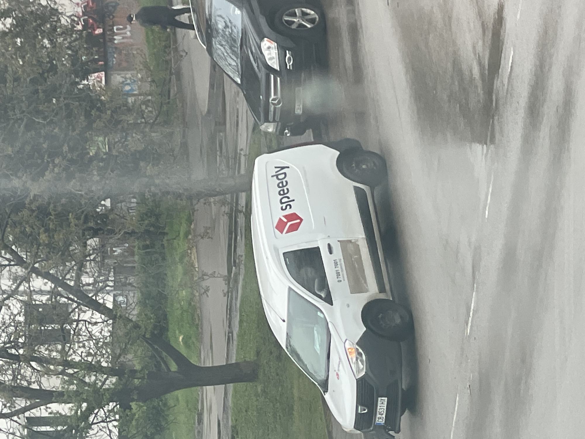 The image displays an outdoor street scene in Sofia, Bulgaria, on an overcast day with a wet road surface.
The primary subject is a white delivery van, likely a Dacia, positioned prominently in the mid-ground. The van belongs to the "Speedy" delivery service, as indicated by the red "Speedy" logo and text on its side. Also visible on the van's side is a phone number, "0 7001 7001". The front license plate, "CB 4531 HX," confirms its registration in Sofia. A person is visible in the driver's seat.
Behind and to the left of the van, a black Mercedes-Benz SUV or car is partially visible. A person dressed in dark clothing is seen walking or standing behind this black vehicle.
The asphalt road is visibly wet, suggesting recent rain or damp conditions. A plume of smoke or mist rises from the area near the front of the black car and the adjacent trees, possibly exhaust or steam from the wet ground.
The background features trees with predominantly bare branches, hinting at late autumn, winter, or early spring. Beyond the trees, some faint structures and a wall with visible graffiti, including the letters "MC," can be discerned. The overall lighting is subdued, typical of a cloudy daytime.