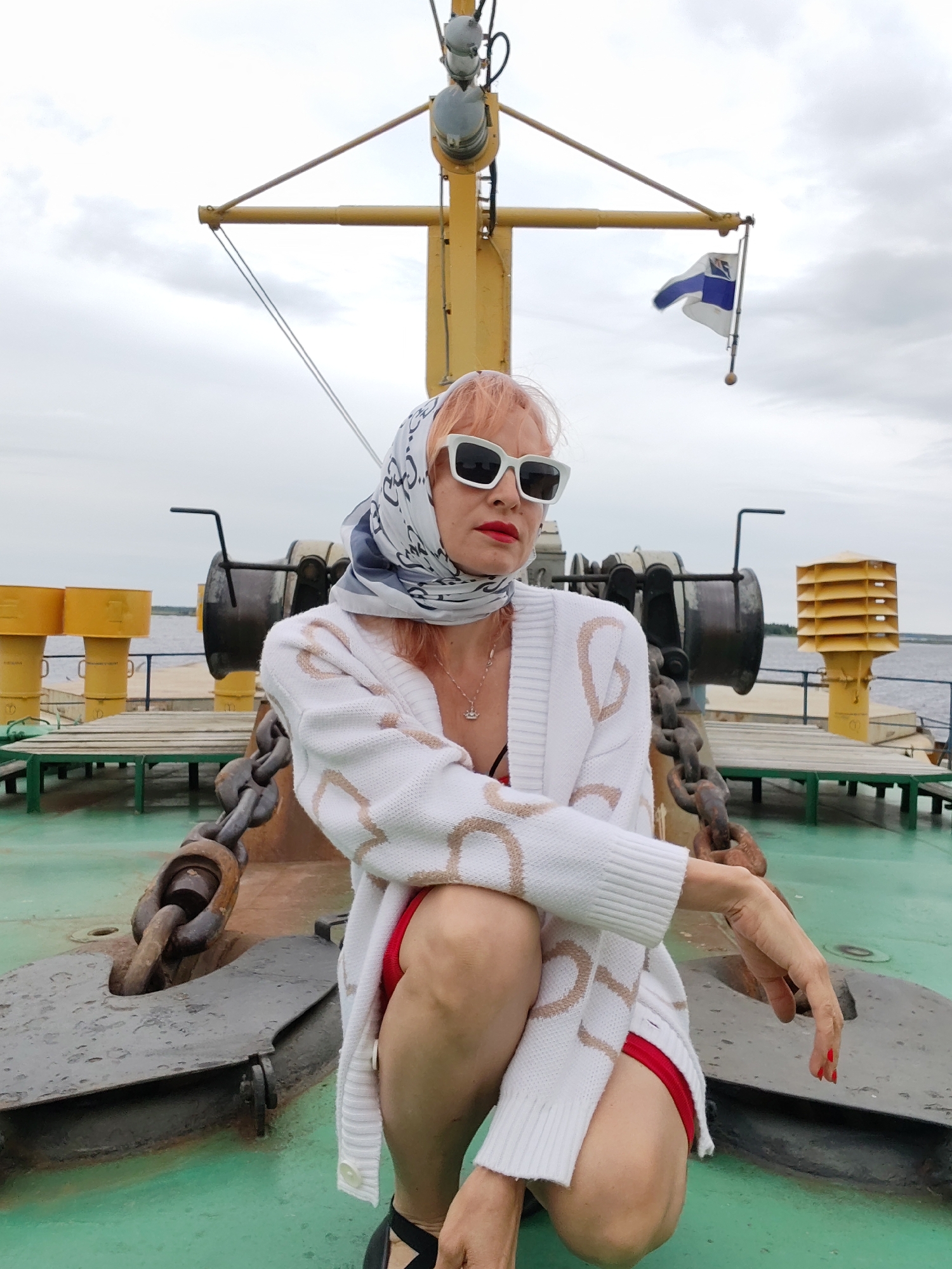 A woman with orange hair, wearing white-rimmed sunglasses, a white patterned cardigan, and a red garment underneath, is posing on the deck of a boat. She is wearing a patterned headscarf. Her makeup includes red lipstick. The boat appears to be a ferry or cargo ship, with heavy anchor chains and metal machinery visible on the deck. Yellow cylindrical objects are present in the background. A flag with blue and white sections is flying on a mast. The weather appears to be overcast, with a cloudy sky. The overall mood is somewhat posed and casual. The time of day seems to be daytime. The location is Novokemskii, Russia.