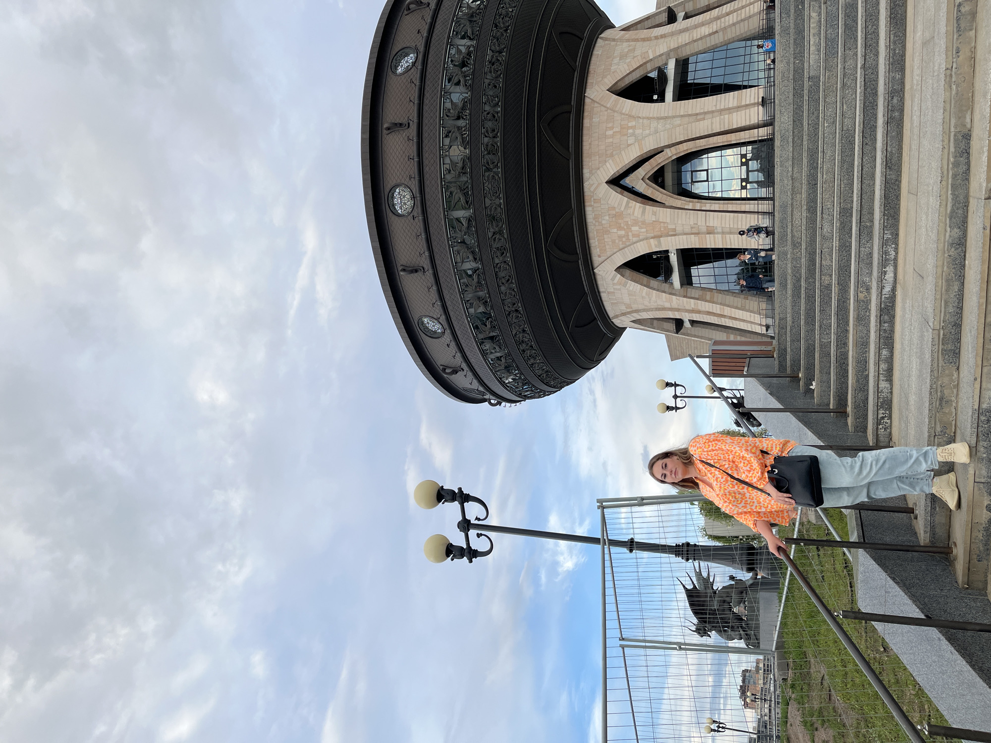 A young woman is posing on a staircase in front of a large, ornate building with arched windows. She is wearing a bright orange and white patterned shirt, light blue jeans, and light-colored boots. She has a black handbag slung over her shoulder. Her pose suggests she is enjoying the moment, with a slight smile and her gaze directed at the camera.
The building behind her appears to be made of stone and metal, with intricate decorative elements. Above the building, the sky is cloudy but bright, suggesting it is daytime. Streetlights are visible in the foreground, and a metal fence runs along the side of the stairs, indicating some ongoing construction or renovation. In the background, beneath the fence, a statue of what appears to be a dragon can be seen. The overall setting is that of an urban environment, with the architecture suggesting a place of cultural or historical significance. The image is oriented vertically, with the ground appearing on the right side of the frame. The specific location is Kazan, Russia.