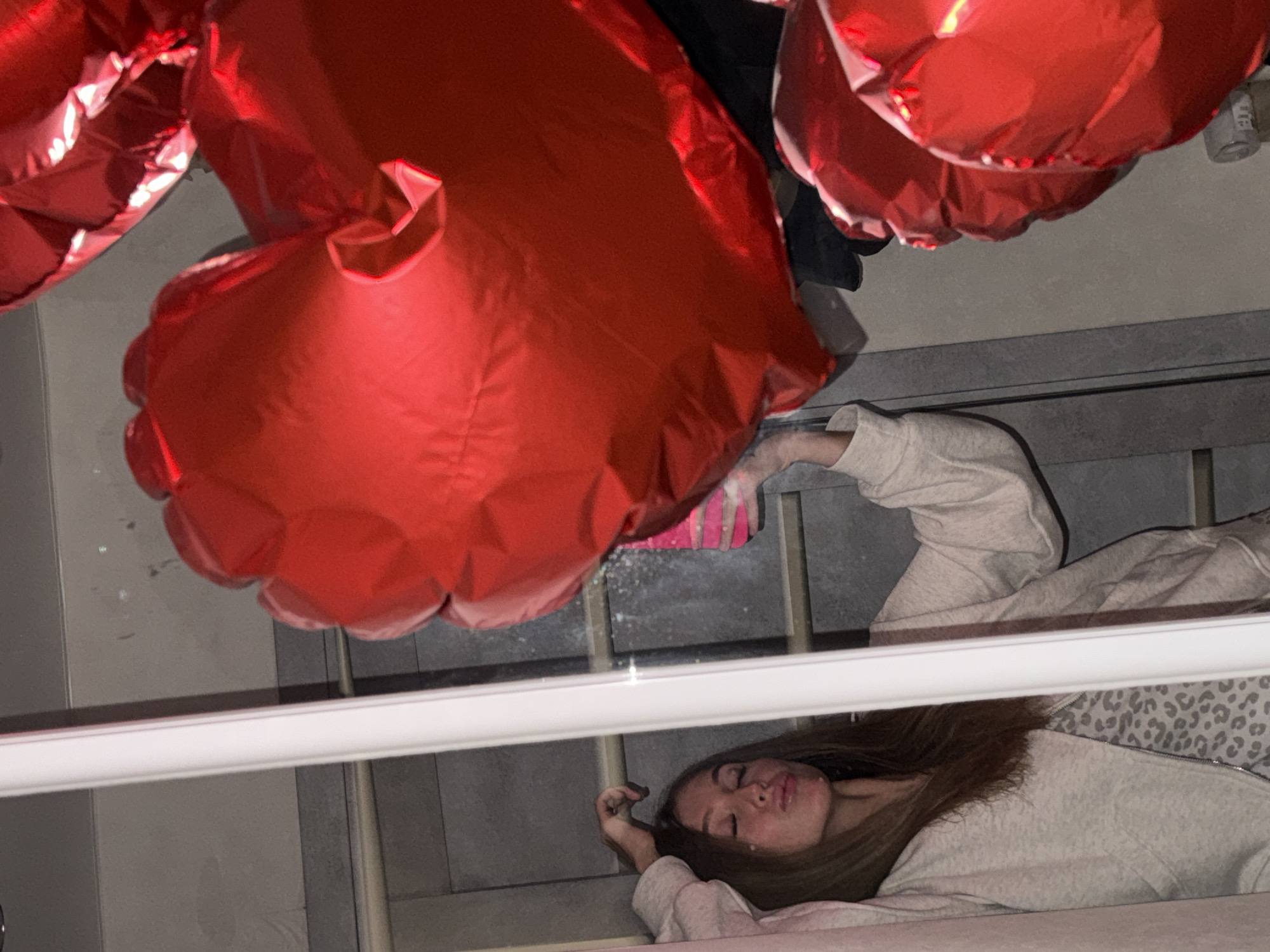 A woman lies on a staircase, seemingly asleep or unconscious. Her head is near the bottom steps, while her body extends upward along the staircase. She is wearing a light gray sweatshirt with a leopard print pattern on the inside of the collar. Above her are several large, red, heart-shaped balloons. A white railing runs horizontally across the middle of the frame. The background consists of the gray concrete walls of the staircase. There is a pink object near the balloons but its not clear what it is. The image is taken from an unusual angle, appearing to be a reflection or captured through a glass surface. There is no visible evidence in the image to confirm the location is Chaikovsky, Russia.