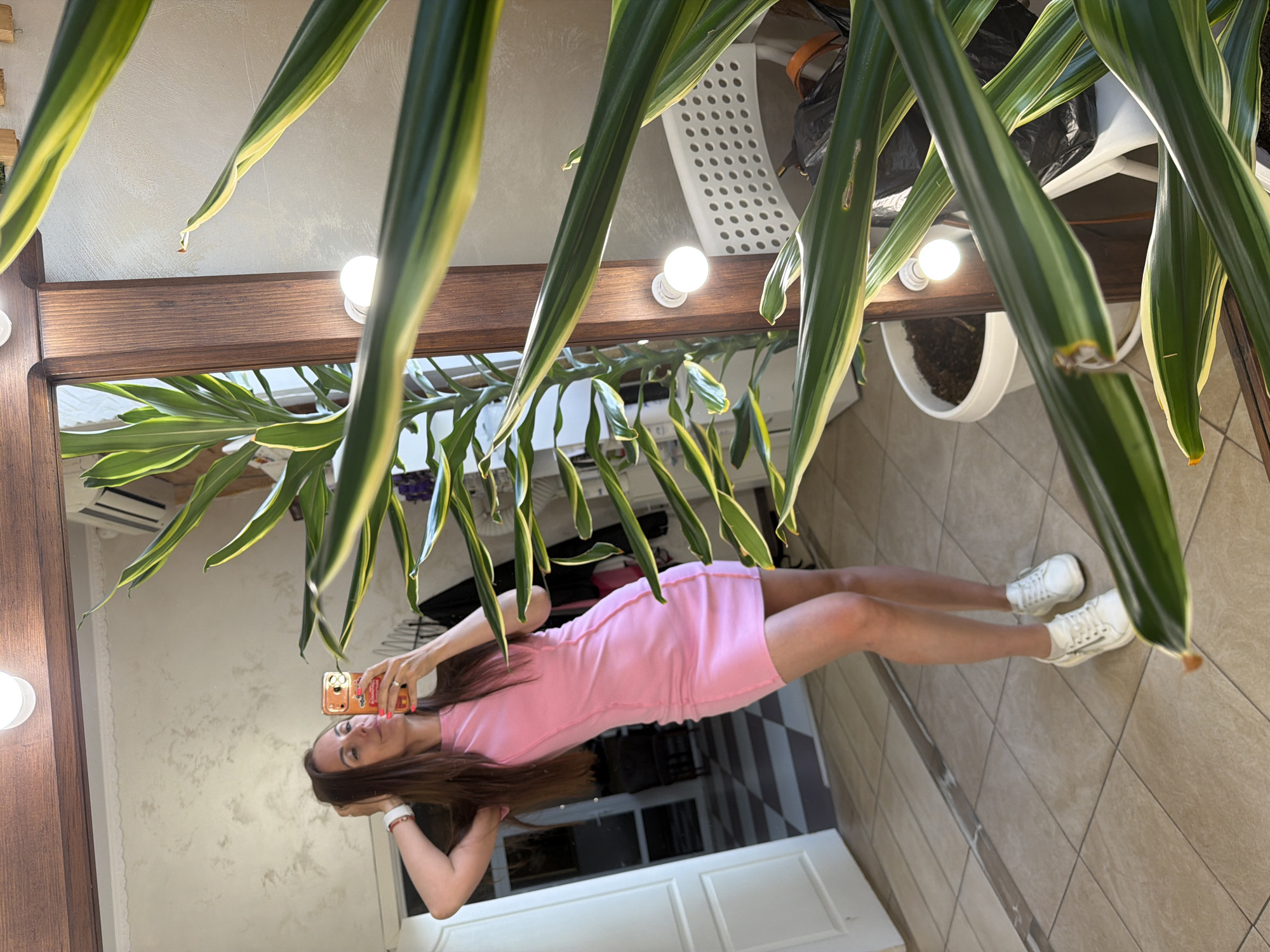 A woman in a pink dress is taking a selfie in front of a mirror. The mirror is surrounded by lights and a wooden frame. There is a large potted plant hanging above the mirror, partially obscuring the view. The background shows a tiled floor and a white cabinet or counter. There is no indication of the location being in Birobidzhan, Russia.