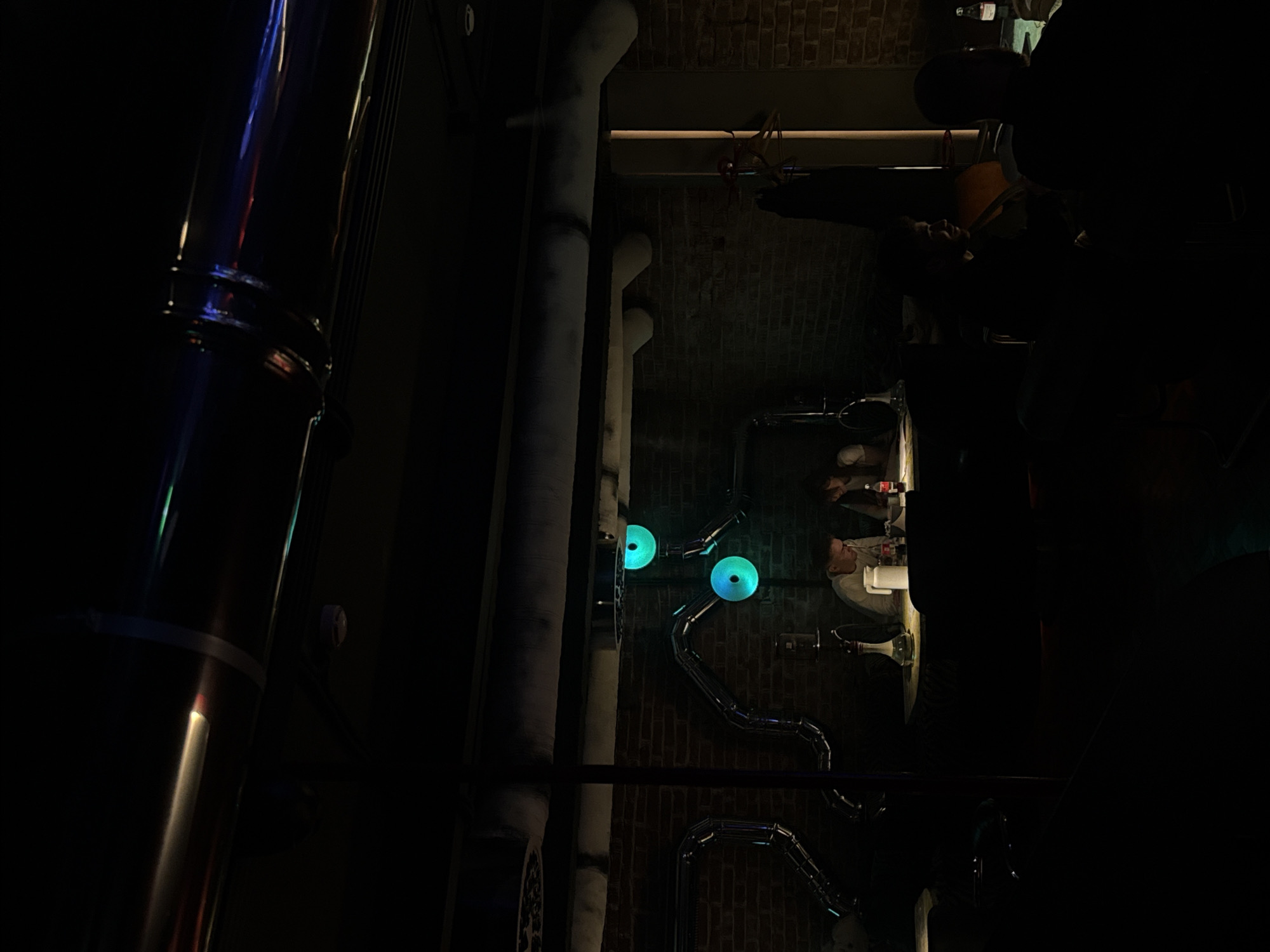 A vertical shot depicts a dimly lit interior of a facility in Moscow, Russia. The scene is dominated by various industrial elements, including several pipes. Some pipes are insulated with white material and run vertically against a brick wall, while others are uninsulated and serpentine, featuring two glowing blue circular lights. In the background, a partially obscured figure appears to be standing at a counter or sink, possibly engaged in some activity, although the darkness makes details unclear. The overall atmosphere is shadowy, and the specific nature of the facility is not immediately apparent from the image.