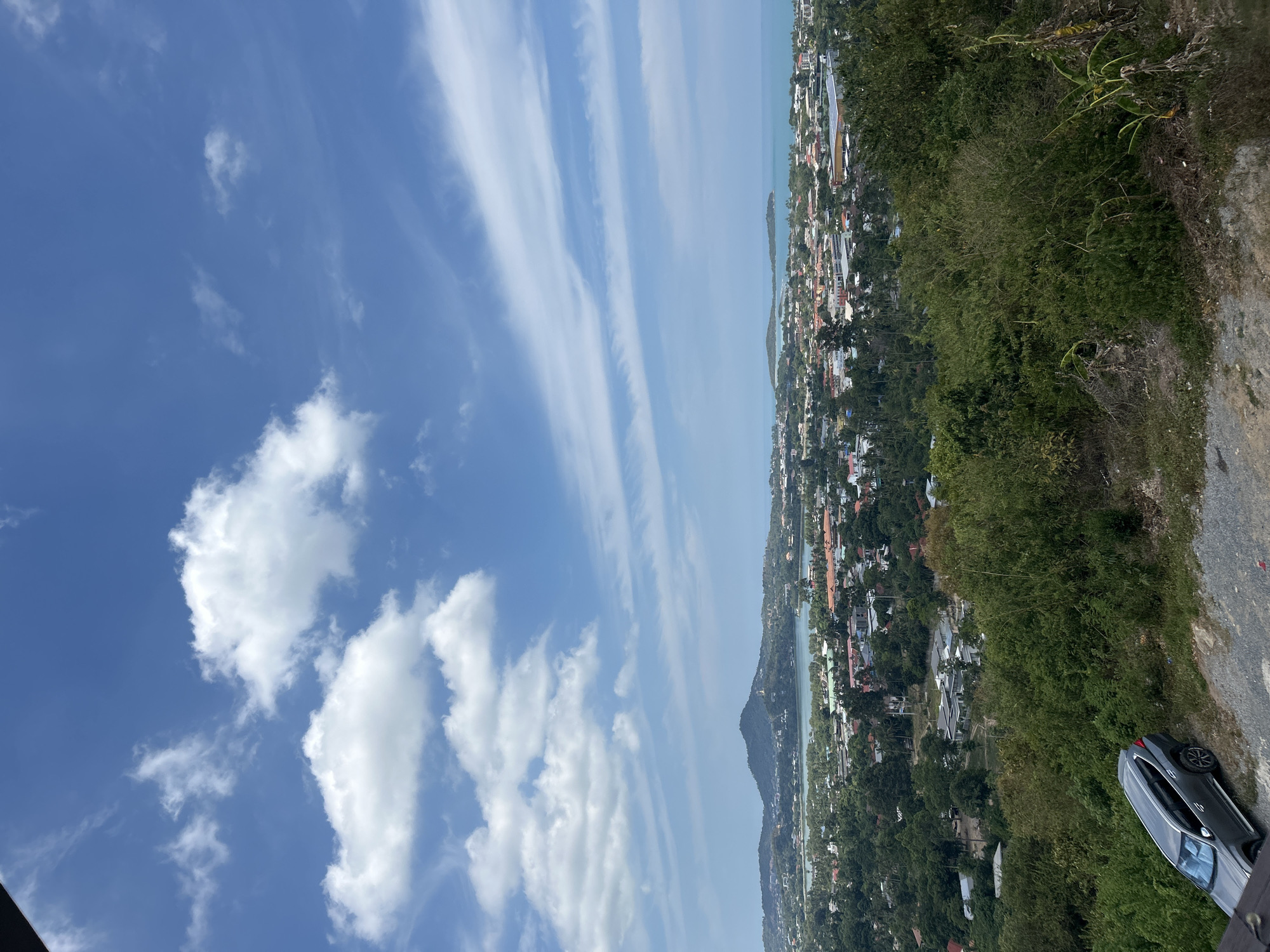 The image depicts a panoramic view of Ko Samui District, Thailand, from an elevated vantage point. The foreground is dominated by dense green foliage and a gray SUV parked on a gravelly incline. Beyond this, a sprawling cityscape unfolds, characterized by a multitude of buildings with orange and red tiled roofs interspersed with lush green trees. A body of water, likely a lake or bay, is visible in the mid-ground, with a prominent mountain rising in the background. The horizon is defined by the turquoise ocean, meeting a bright blue sky dotted with white and wispy clouds. The overall scene suggests a sunny day with clear visibility, capturing the tropical landscape and urban development of Ko Samui.
