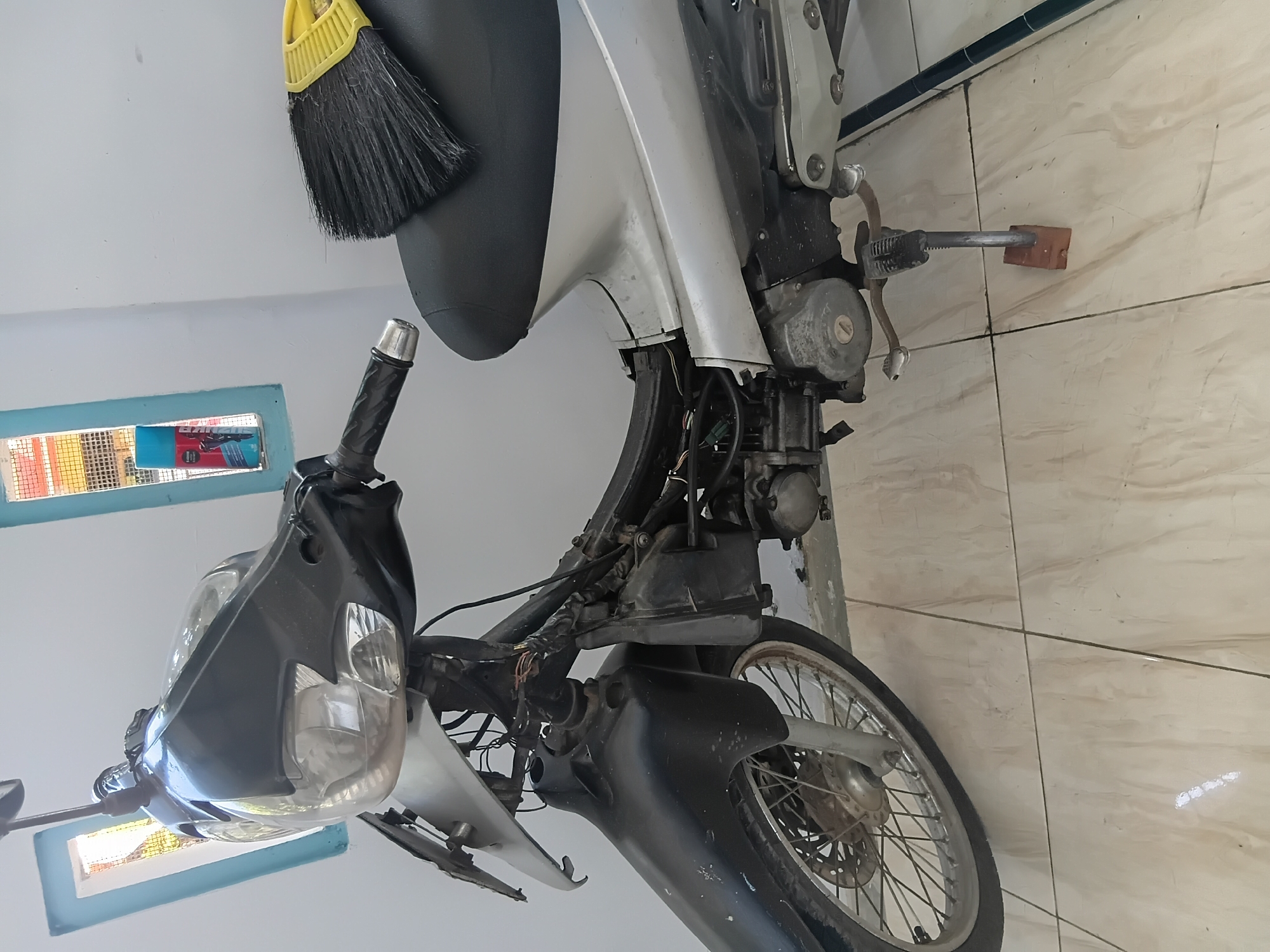 A motorcycle is parked on a tiled floor inside a structure with white walls. The motorcycle is silver and black, with exposed wiring near the handlebars. A black broom with a yellow handle rests on the seat. Behind the motorcycle, there are two windows with blue frames, one of which contains a can of spray paint. The scene appears to be in Tanggul Wetan, Indonesia.