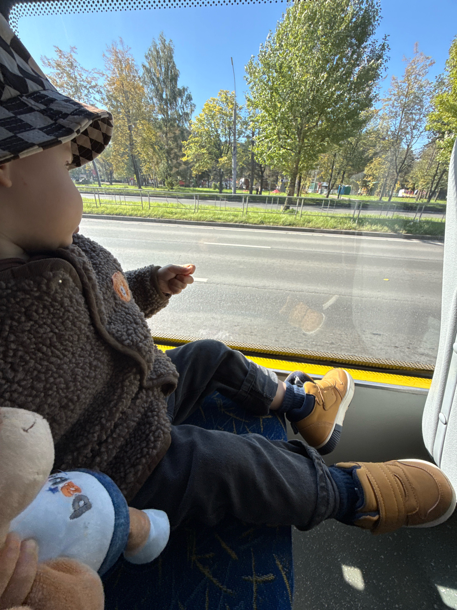 A young child wearing a checkered bucket hat and a brown fuzzy jacket sits on a bus seat, looking out the window. The child's legs are extended, and they are wearing dark pants and brown sneakers. A stuffed animal rests on their lap. Outside the window, trees with yellowing leaves line a street with a road and sidewalk. The sky is clear and blue. The scene appears to be in Rusanovo, Russia, during autumn.