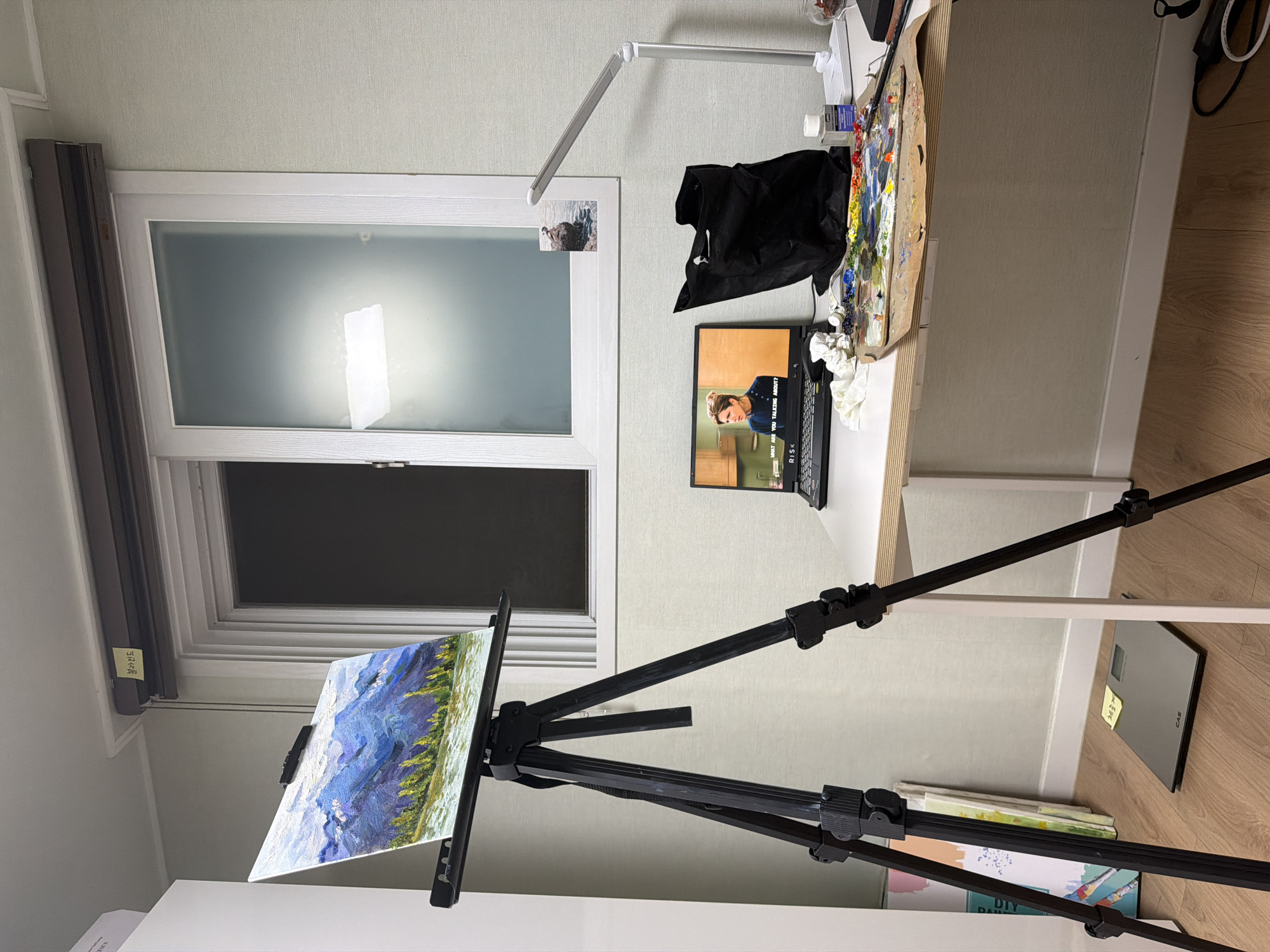 The image depicts an art studio setup in an indoor residential space, likely an apartment, in Dongducheon-si, South Korea. The room is illuminated by artificial light from a desk lamp and the light from a laptop screen. A landscape painting of mountains and trees is displayed on a tripod easel. To the right of the easel, a desk holds a laptop displaying a scene from a television show or movie, a black tote bag, a palette with various colors of paint, brushes, and a bottle of liquid. A window with frosted glass is visible in the background. On the floor, a digital scale with a yellow sticky note is present, along with several canvases leaning against a wall. The overall scene suggests a creative space for painting, possibly with a viewer or reference material displayed on the laptop.