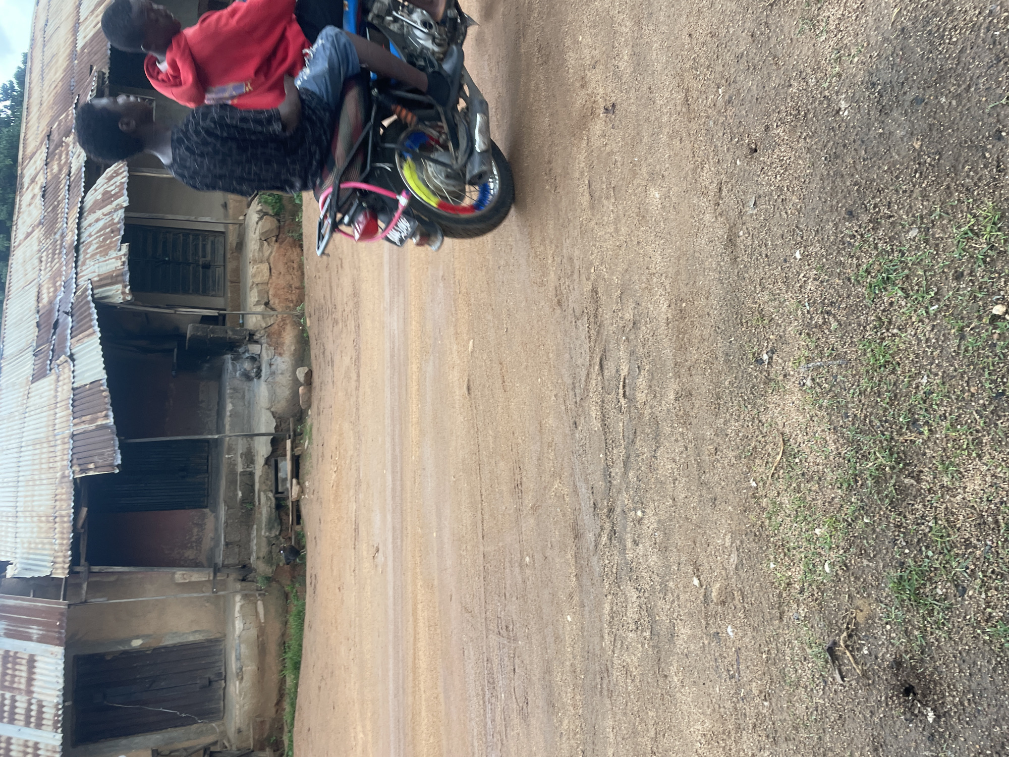In Gwong, Nigeria, two young Black males are riding a motorcycle on a dirt road in front of a dilapidated building. The building has a corrugated metal roof, wooden doors, and a rough, earthy facade. One of the males is wearing a black t-shirt with a geometric pattern, and the other is wearing a red hooded jacket. The motorcycle is blue and black, with a red taillight and colorful spokes on the rear wheel. The road is unpaved and appears to be dusty and possibly damp in places. Some green vegetation is visible near the building and the edge of the road.