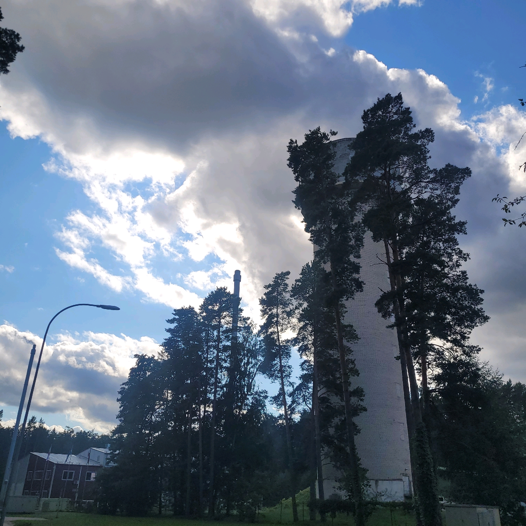 This image depicts a view of a tall, cylindrical white brick structure, likely a silo or a tower, situated within a natural setting of dense pine trees. The structure is partially obscured by the foliage of the trees, with sunlight filtering through the branches. In the foreground and to the left, a dark, curved lamppost is visible against a sky filled with dramatic, dynamic clouds. The clouds are a mix of bright white, suggesting sunlight, and dark gray, indicating potential storminess or heavy cloud cover. Further to the left, a low-rise building with a red facade and a white roof is present, partially visible through the trees. The ground appears to be covered in grass. The location is identified as Jūrmala, Latvia, suggesting this scene is part of the coastal resort city's landscape. There are no people or animals visible in the image. The scene is static, focusing on the architectural structure, the surrounding vegetation, and the atmospheric conditions of the sky.