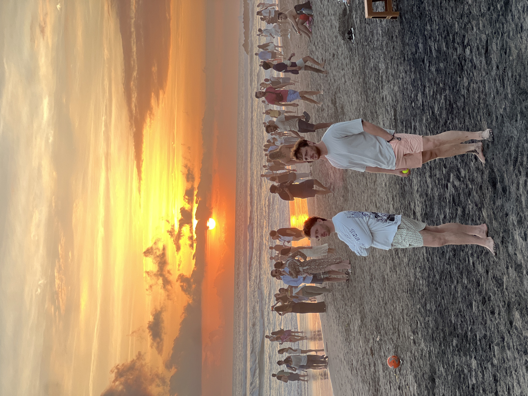 The image depicts a beach scene in North Kuta, Indonesia, during sunset. The sky is filled with vibrant orange and yellow hues, with scattered clouds. The sun, a bright orb, is setting over the ocean, casting a warm glow on the water and the wet sand. A crowd of people are gathered on the beach, some wading in the shallow water, others standing on the sand.
In the foreground, two young men are visible. The man on the left is wearing a white long-sleeved shirt with a graphic print and patterned shorts. He is standing with his arms crossed, looking towards the ocean. To his right, another man is wearing a light gray t-shirt and pink shorts. He has his hands in his pockets and is looking away from the camera.
Further back, a diverse group of people are visible. Some are casually dressed, while others appear to be dressed for an evening out. They are interacting with each other, walking along the beach, or simply observing the sunset. An orange ball is visible on the sand near the man in the white shirt, suggesting a game or activity. The sand appears dark and wet, indicative of the tide. The overall atmosphere suggests a relaxed and social gathering.