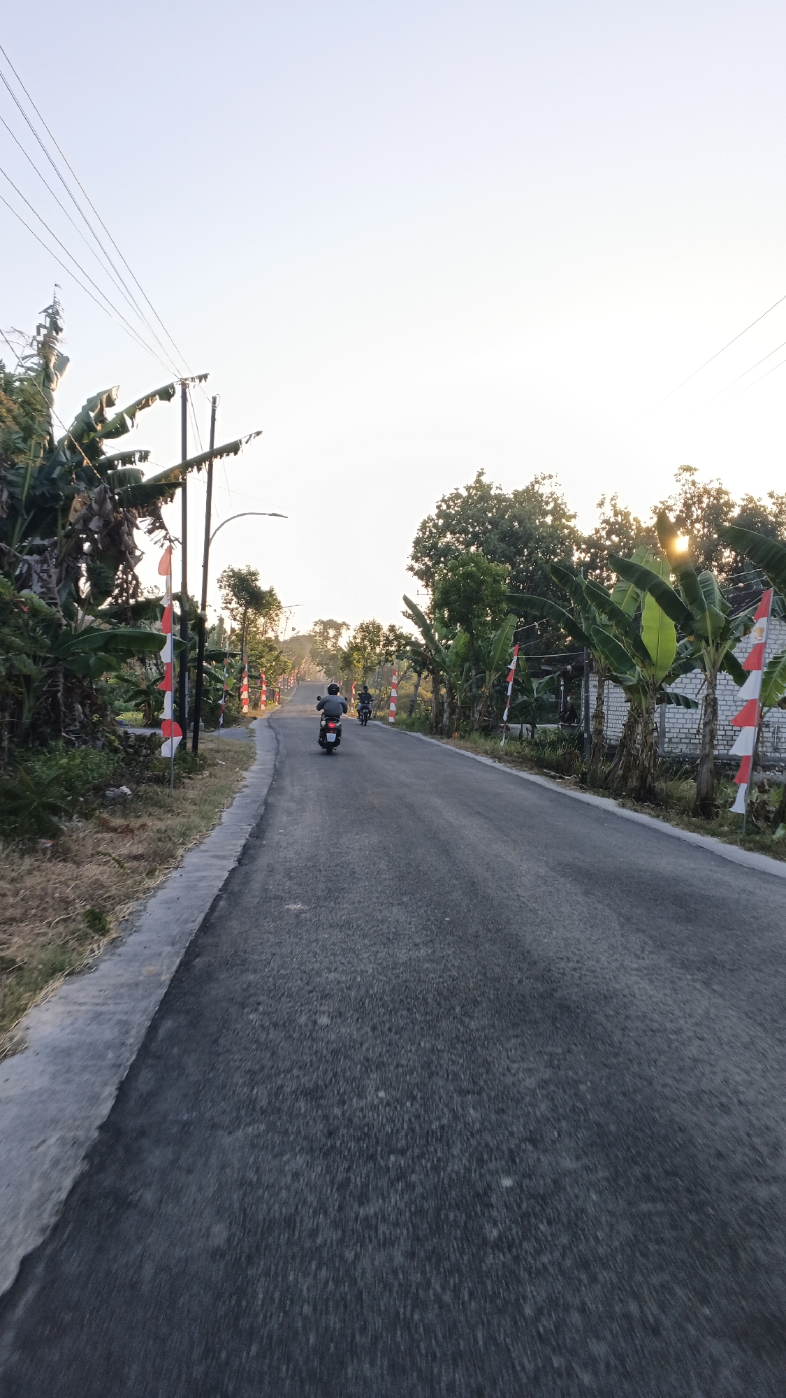The image depicts a rural road in the city of Soko, Indonesia, during what appears to be either sunrise or sunset. The road is paved with asphalt and is flanked by lush vegetation, including banana trees. Red and white Indonesian flags are prominently displayed along the roadside, suggesting a local celebration or commemoration. Two individuals are seen riding motorcycles on the road. The one in the foreground is facing away from the camera, while the second motorcyclist is further ahead and also facing away. The sky is bright with sunlight filtering through the trees, creating a warm, ambient light. Utility poles and power lines are visible on the left side of the road. The overall scene conveys a sense of peaceful rural life in Indonesia.
