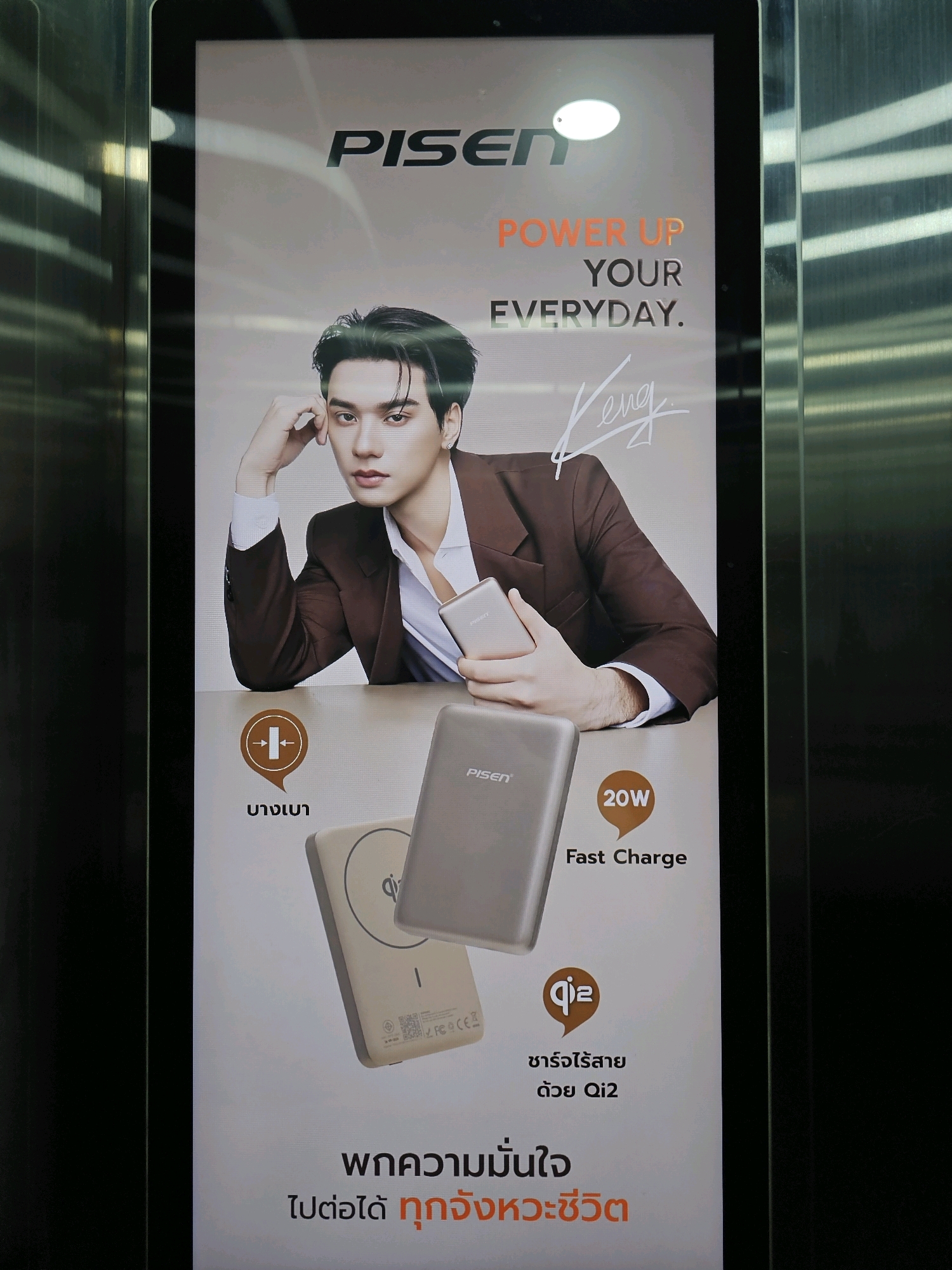 The image is an advertisement for PISEN portable chargers, displayed on a digital screen likely located in a public space in Bangkok, Thailand, possibly an elevator or a mall. The advertisement features a young Asian man in a brown suit holding two PISEN chargers. One charger is white and rectangular with the "Qi2" logo and text in Thai indicating wireless charging. The other charger is silver and appears to be a power bank, labeled "20W Fast Charge". The advertisement also includes the PISEN logo and the slogan "POWER UP YOUR EVERYDAY." The background is a gradient of light gray and white. The framing of the advertisement suggests it is a vertical digital display.