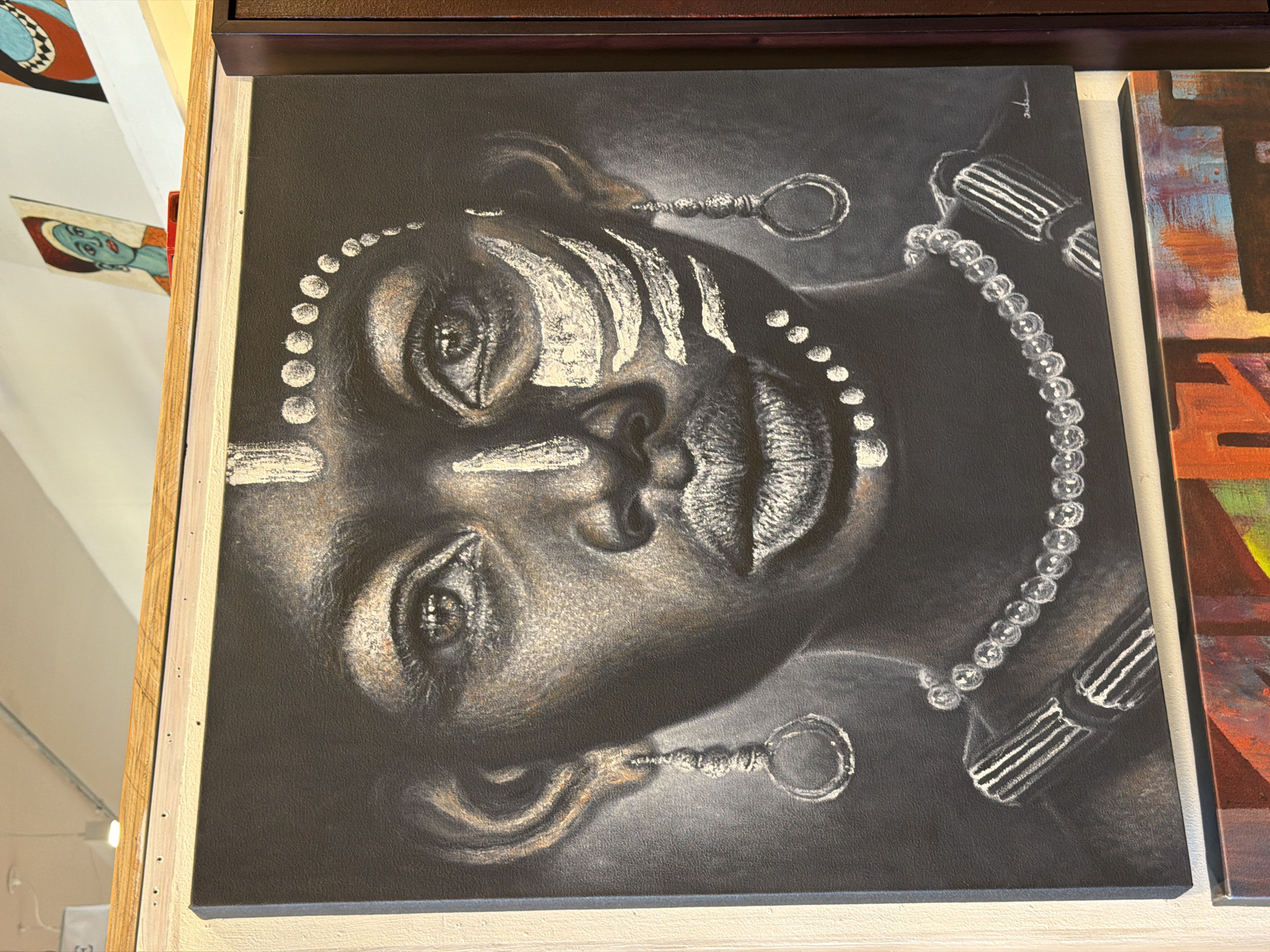 The image features a close-up of a striking portrait painting, displayed vertically, but depicting a person's face tilted about 90 degrees to the left. The artwork is characterized by a monochrome palette, using dark greys and blacks as a base, with prominent white and subtle light brown/gold highlights, suggesting a medium like charcoal, chalk, or textured paint on canvas.
The subject's face is adorned with intricate white markings: a series of dots extends across the forehead, down the side of the face, and forms a line resembling a necklace. Bold, stylized white stripes are painted on the forehead and cheeks, evocative of traditional body or tribal art. The person has open, expressive eyes and full, slightly parted lips. Large circular earrings with textured cylindrical elements are visible on both ears, and a prominent strand of white, pearl-like beads graces the neck and lower face area. On the right side, a cylindrical armband or bracelet with striped white patterns is depicted. A faint signature, possibly "Tepo," is visible in white script in the top right corner of the painting.
The painting is part of an indoor art display, likely in a gallery, studio, or market. It has a light wooden border on its left and bottom edges. To its right, a portion of another colorful, abstract painting with geometric shapes in reds, oranges, and blues is visible. Further in the background, other artworks can be seen, including one with a teal-skinned face. The bright lighting suggests an indoor setting with natural illumination, creating an exhibition ambiance. No specific activity is taking place other than the presentation of the art. No visible text beyond the potential artist's signature is discernible. While the location context of Cape Town, South Africa, was provided, the image itself does not contain explicit visual cues to confirm this geographical location.