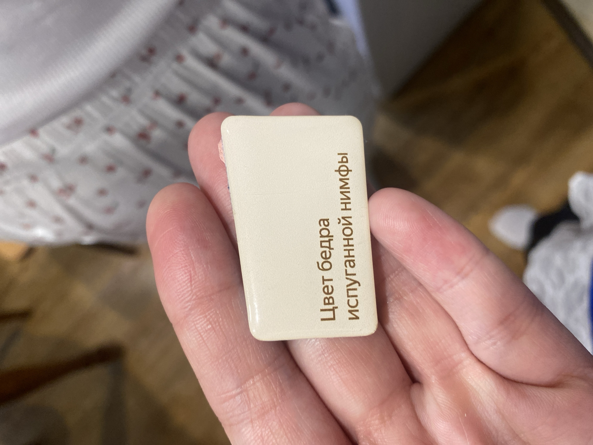 A hand holds a rectangular paint sample card. The card is off-white and has brown Cyrillic text that reads "Цвет бедра испуганной нимфы" (Color of a frightened nymph's thigh). The location is unknown.