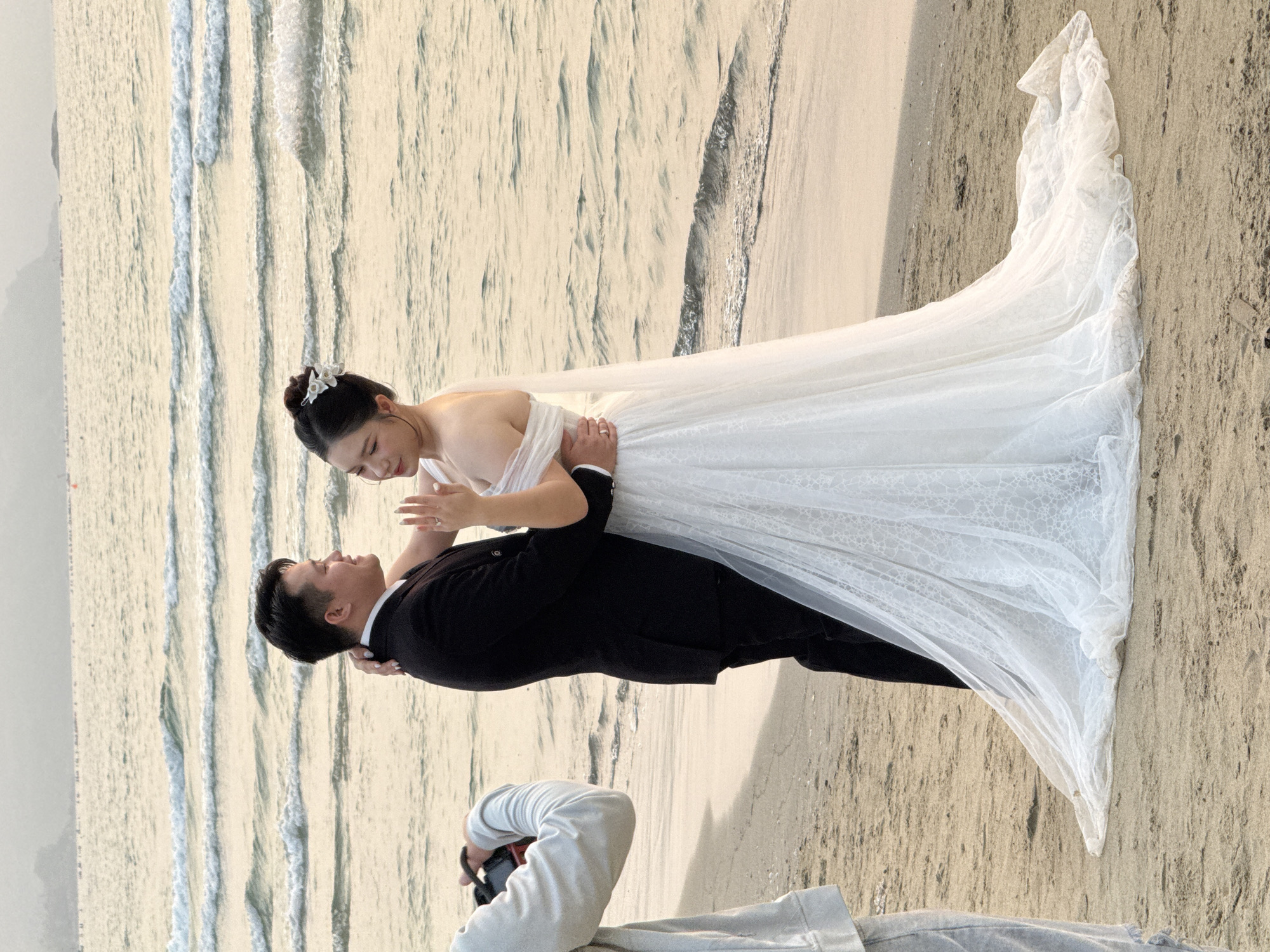 A wedding photoshoot is in progress on a sandy beach with the ocean waves visible in the background. A bride wearing a white wedding dress and a groom in a black suit are the focal point of the scene. The groom is holding the bride in a pose, while a photographer is seen kneeling in the lower portion of the frame, capturing the moment with a camera. The location is likely on a beach in Đà Nẵng, Vietnam.