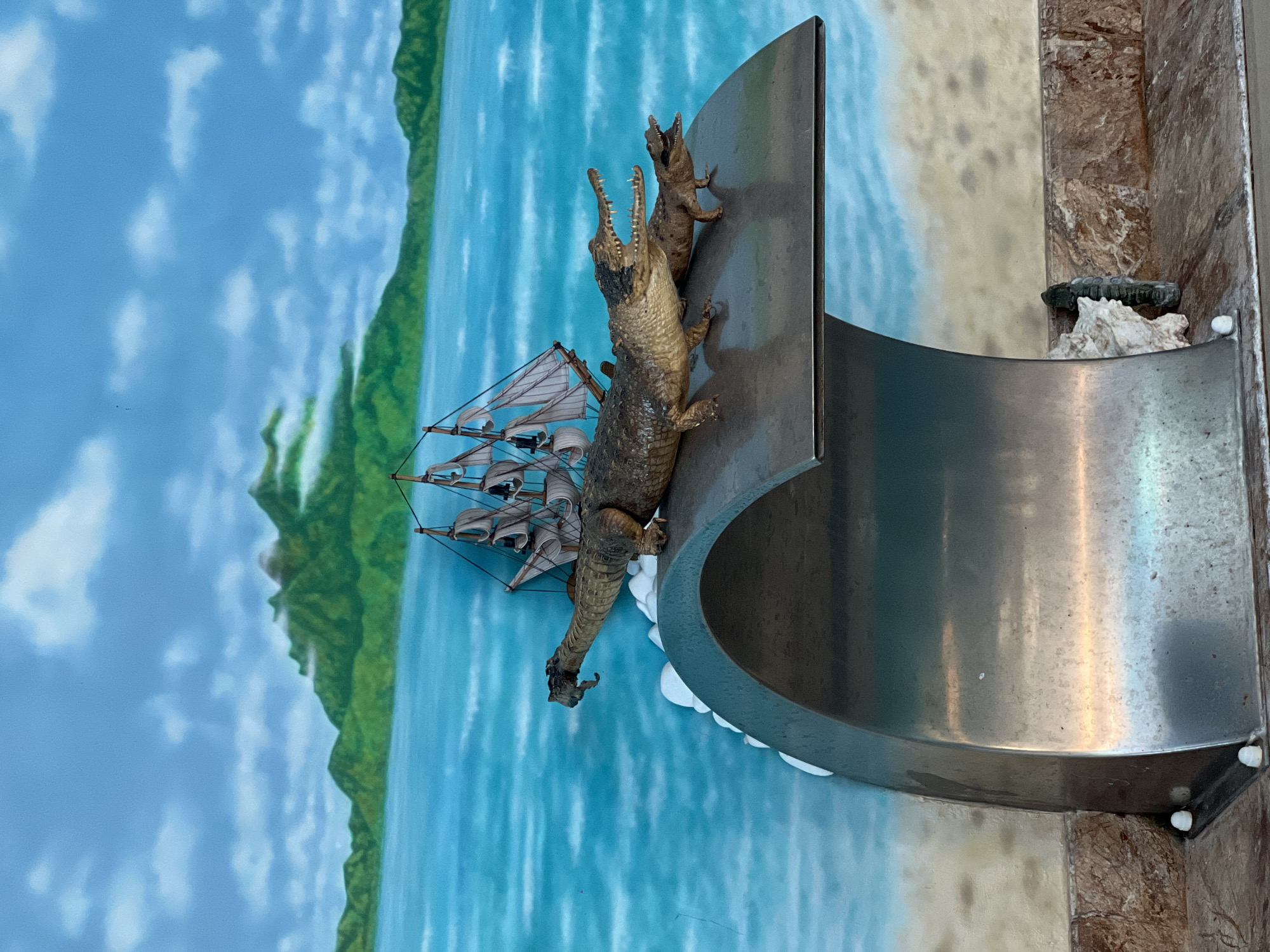 This image depicts a decorative waterfall feature in Bender, Moldova, designed to resemble a tropical seascape. The backdrop is a mural of a blue sky with white clouds, green, misty mountains, and a turquoise ocean. A sailing ship is depicted in the background. In the foreground, resting on the metallic lip of the waterfall, are two realistic toy crocodiles. The larger crocodile is in the foreground, its mouth open, and a smaller crocodile is positioned behind it, also with its mouth open. The waterfall itself is made of polished metal and curves downwards. The surrounding environment includes a sandy-colored wall or floor with visible marble-like patterns. There are no people visible in the image.